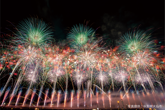 Colorful fireworks erupt in a dark night sky above a crowd, with green, red, and white bursts lighting the scene.