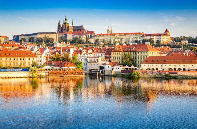 Sunlit European riverside town with red-roofed houses and a historic castle on a hill, reflected in calm water.