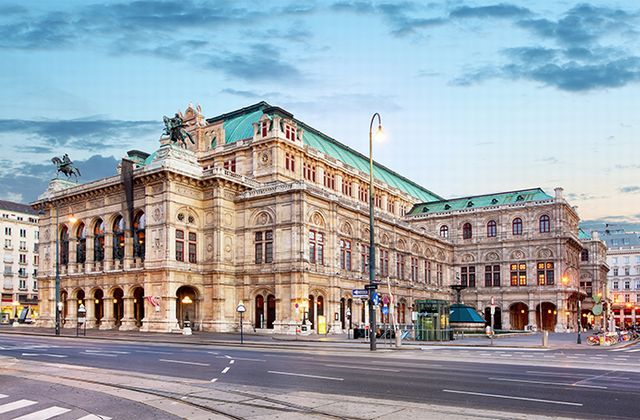 Ornate historic opera house with a green copper roof on a city street at dusk, horses statue visible on the left façade.
