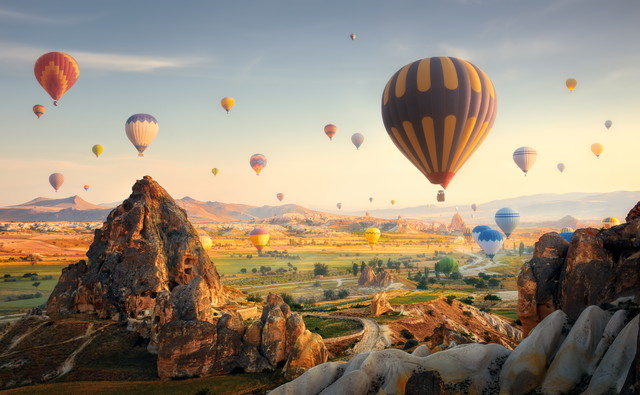 Dozens of colorful hot air balloons floating over a rocky valley at dawn, with a large striped balloon in the foreground and warm sunrise light.