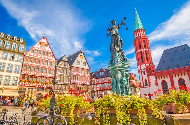 Colorful half-timbered houses surround a plaza with a bronze statue on a pedestal and a flower-filled fountain in the foreground.