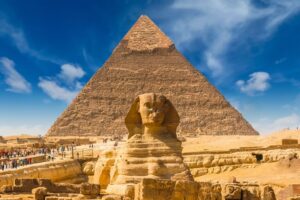 Sphinx statue with the Pyramid of Giza behind, in a desert plaza under a blue sky with visitors nearby.