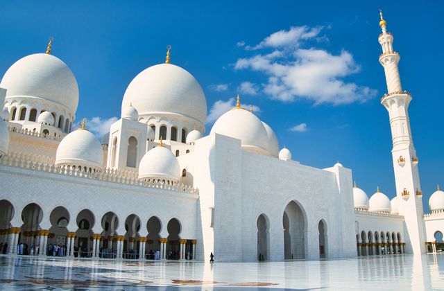 Exterior view of a grand white mosque with large domes and a tall minaret against a bright blue sky, Abu Dhabi