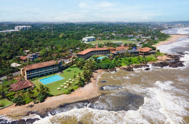 Aerial view of a beachfront resort with a rectangular pool, palm trees, and orange-roofed buildings along a rocky shoreline.