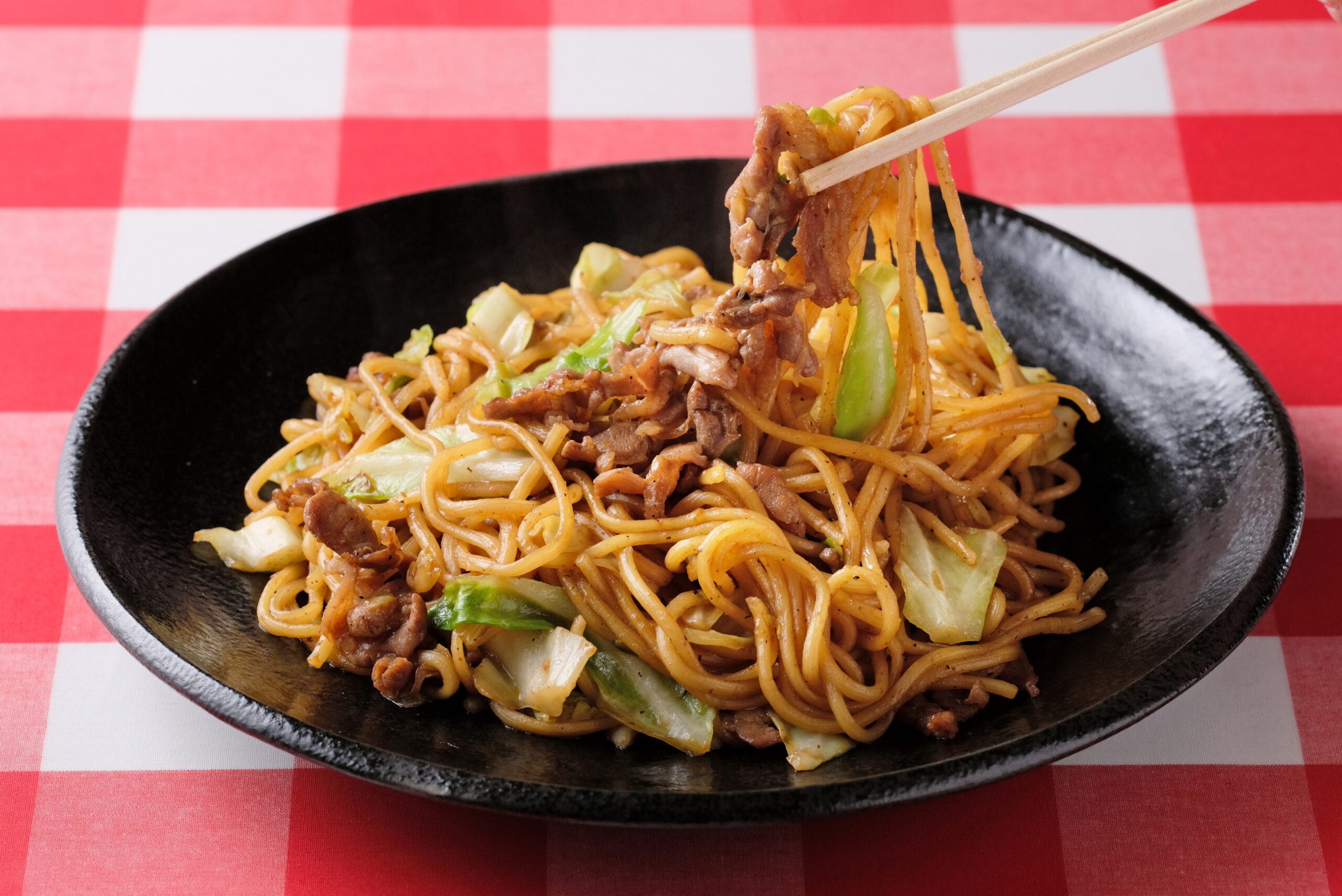 Bowl of stir-fried noodles with beef and cabbage on a red-and-white checkered tablecloth with chopsticks lifting noodles