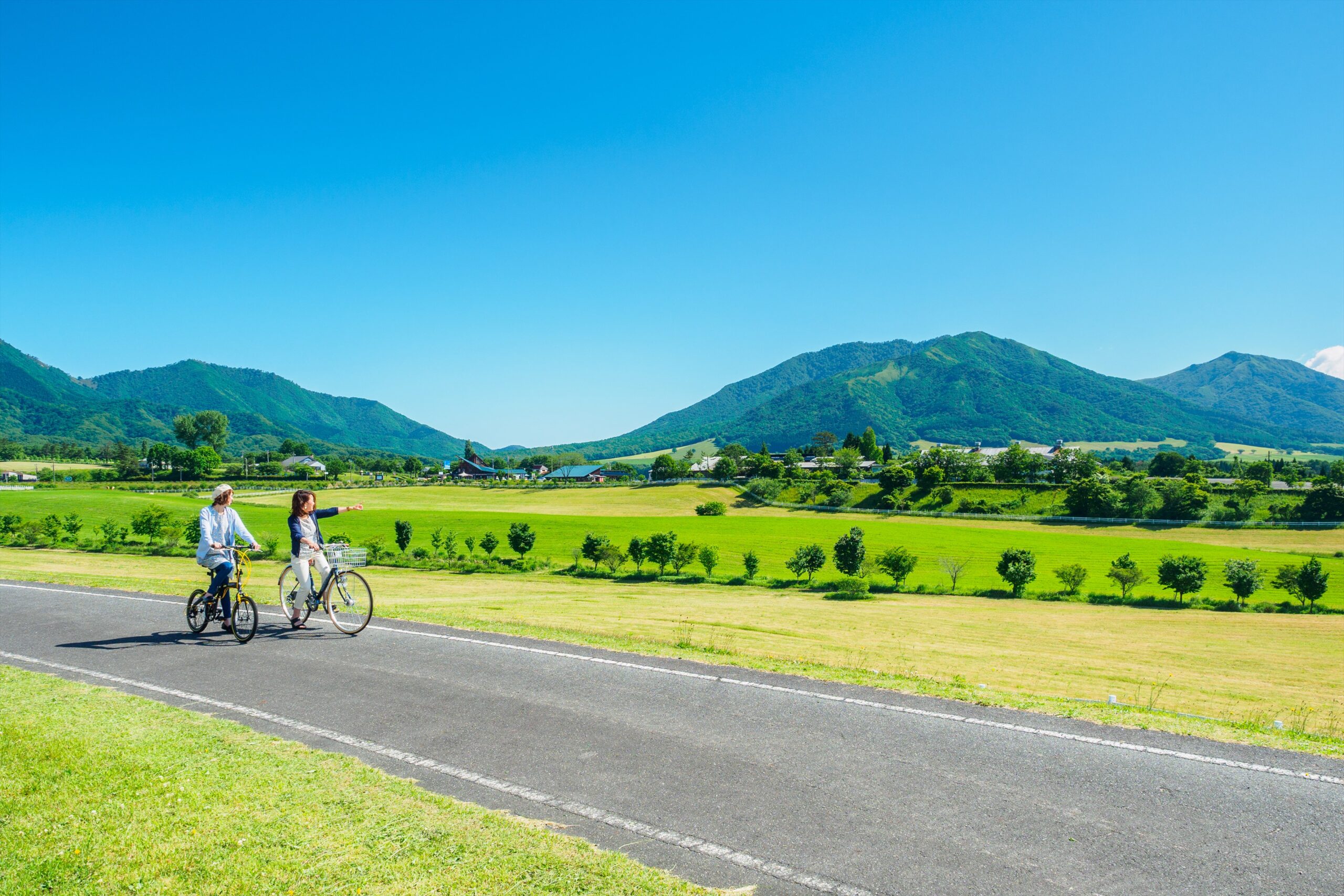 Two people ride bicycles on a paved path through a sunny rural landscape with green fields and distant mountains under a clear blue sky