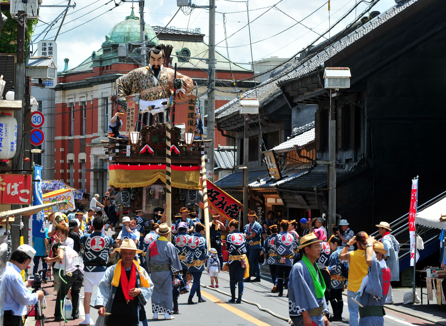 Colorful Japanese street festival with a tall wooden float featuring a giant warrior figure, banners, and gold fringe, surrounded by people in blue and white happi coats.