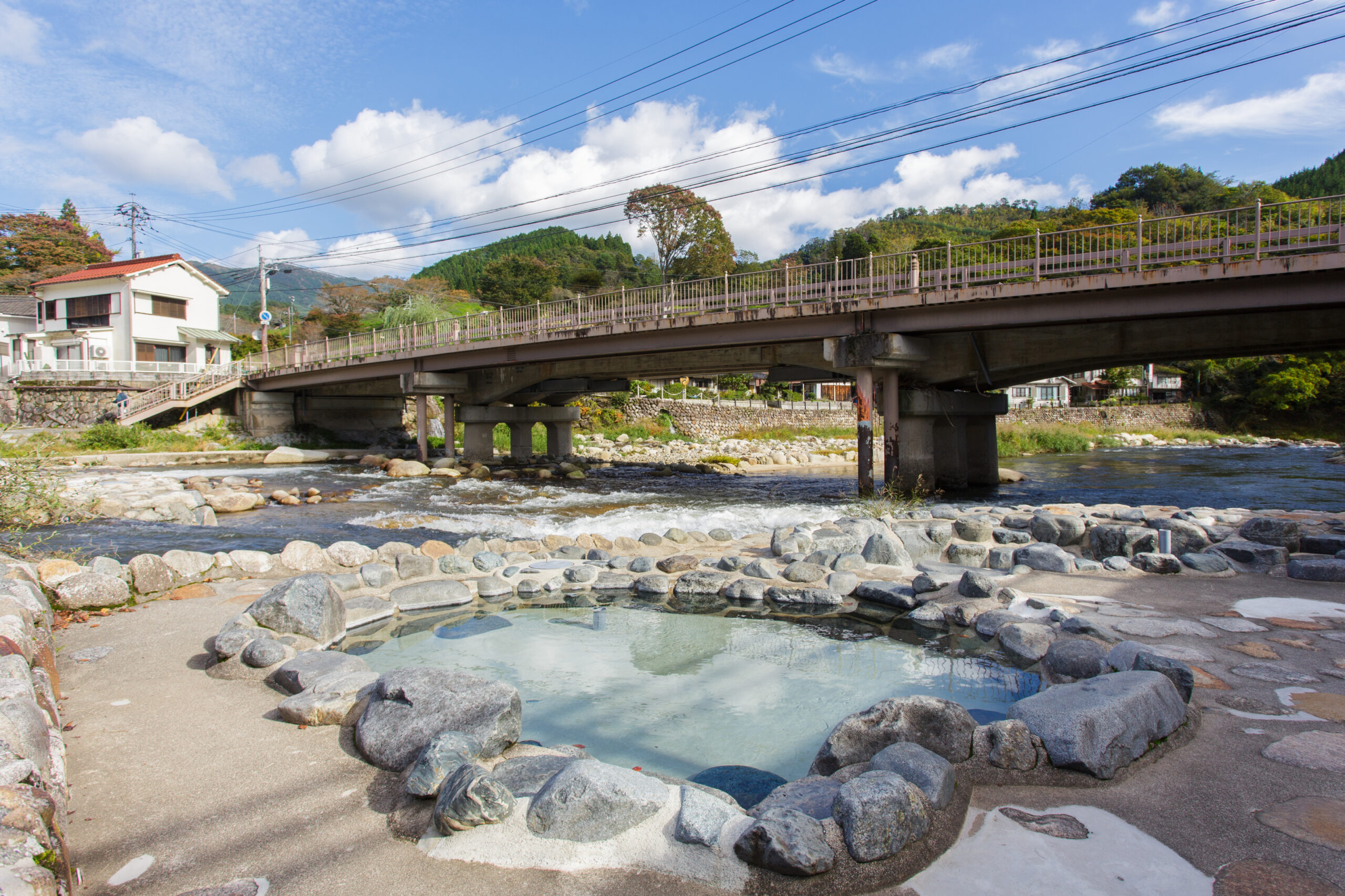 Bridge over a rocky river with a small town on the left and green hills in the background under a blue sky.