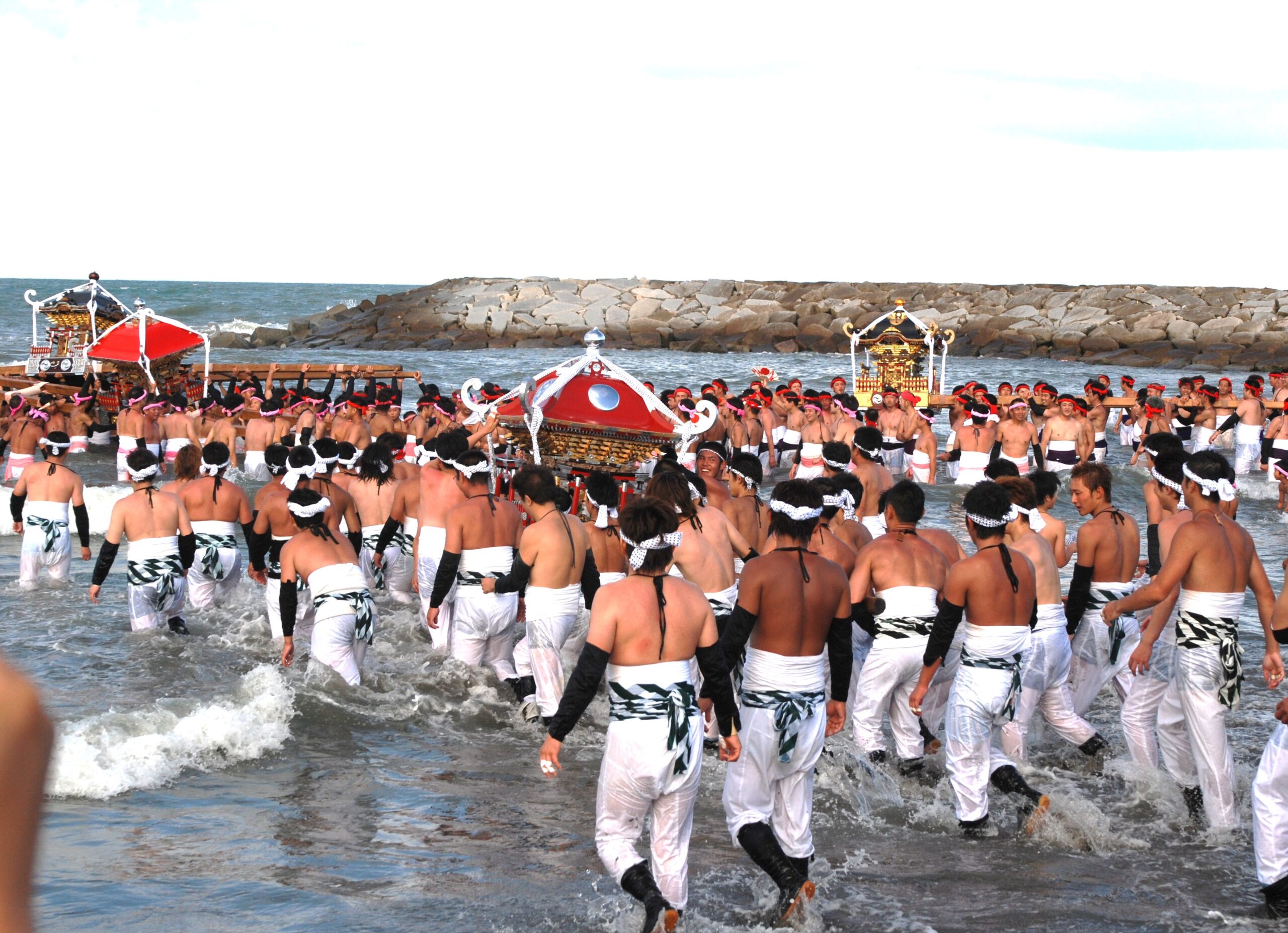 Large crowd of shirtless men wearing white belts and headbands wading into the sea for a festival, carrying ornate portable shrines.
