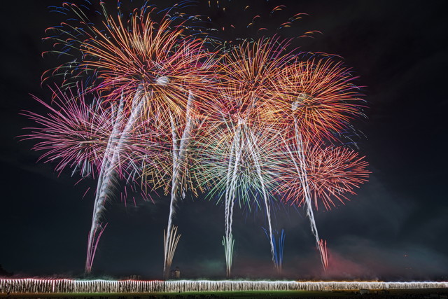 Nighttime fireworks display bursting in red, orange, and blue over a dark field with smoke and a row of ground lights below.