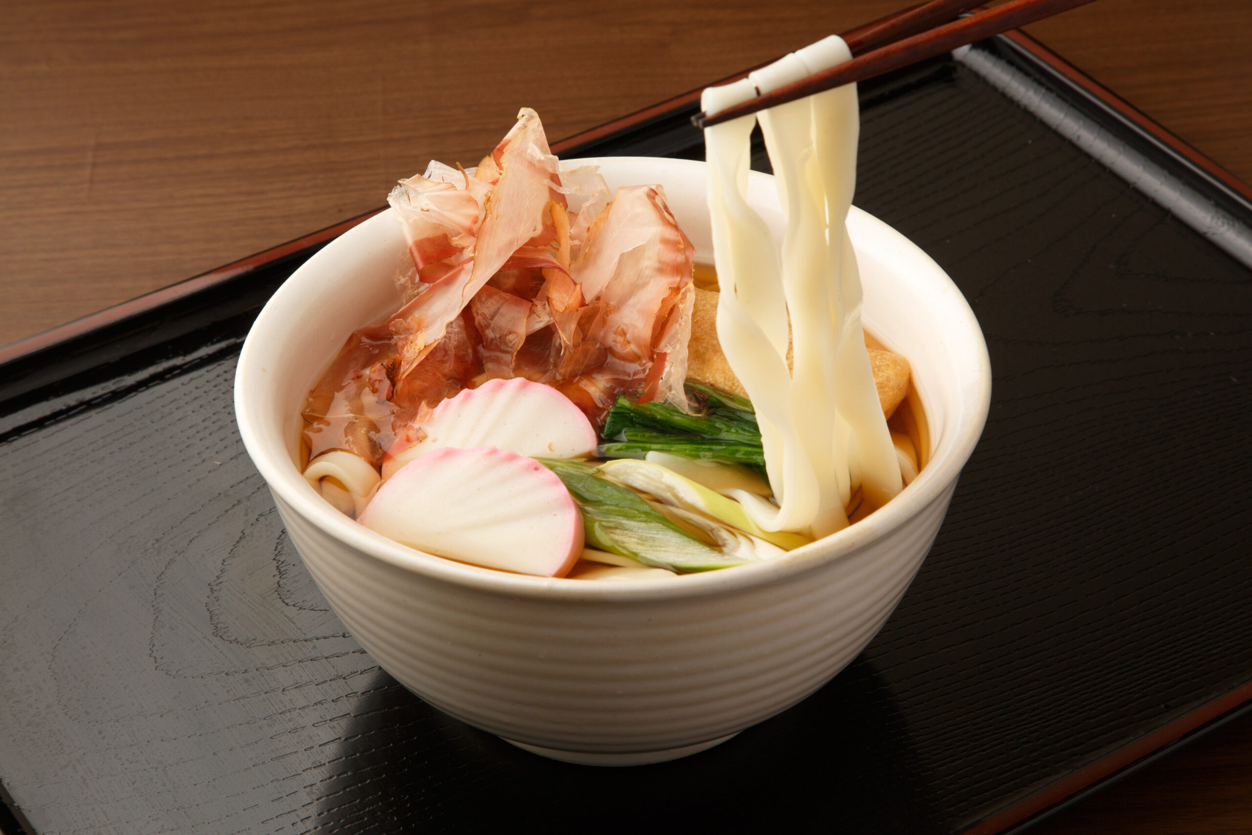 Bowl of udon noodles with sliced fish cake, greens, and bonito flakes on a dark wooden tray.