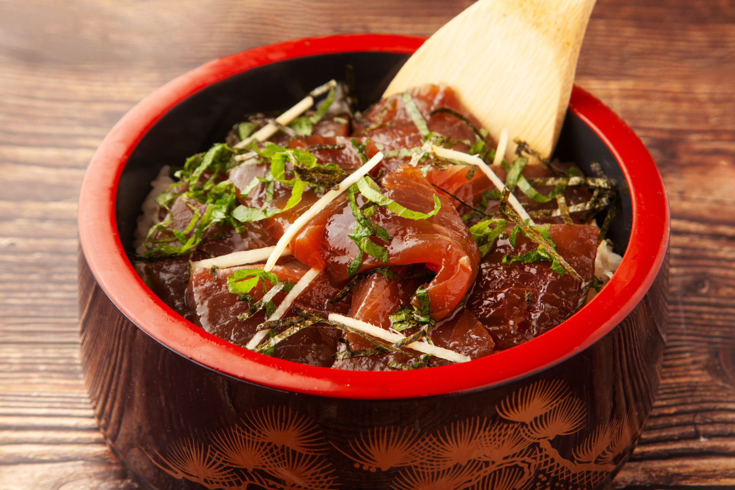 Red-lacquered bowl of sashimi donburi with sliced tuna, shredded seaweed and white garnish over rice, on a wooden table with a wooden spoon welcome image context.