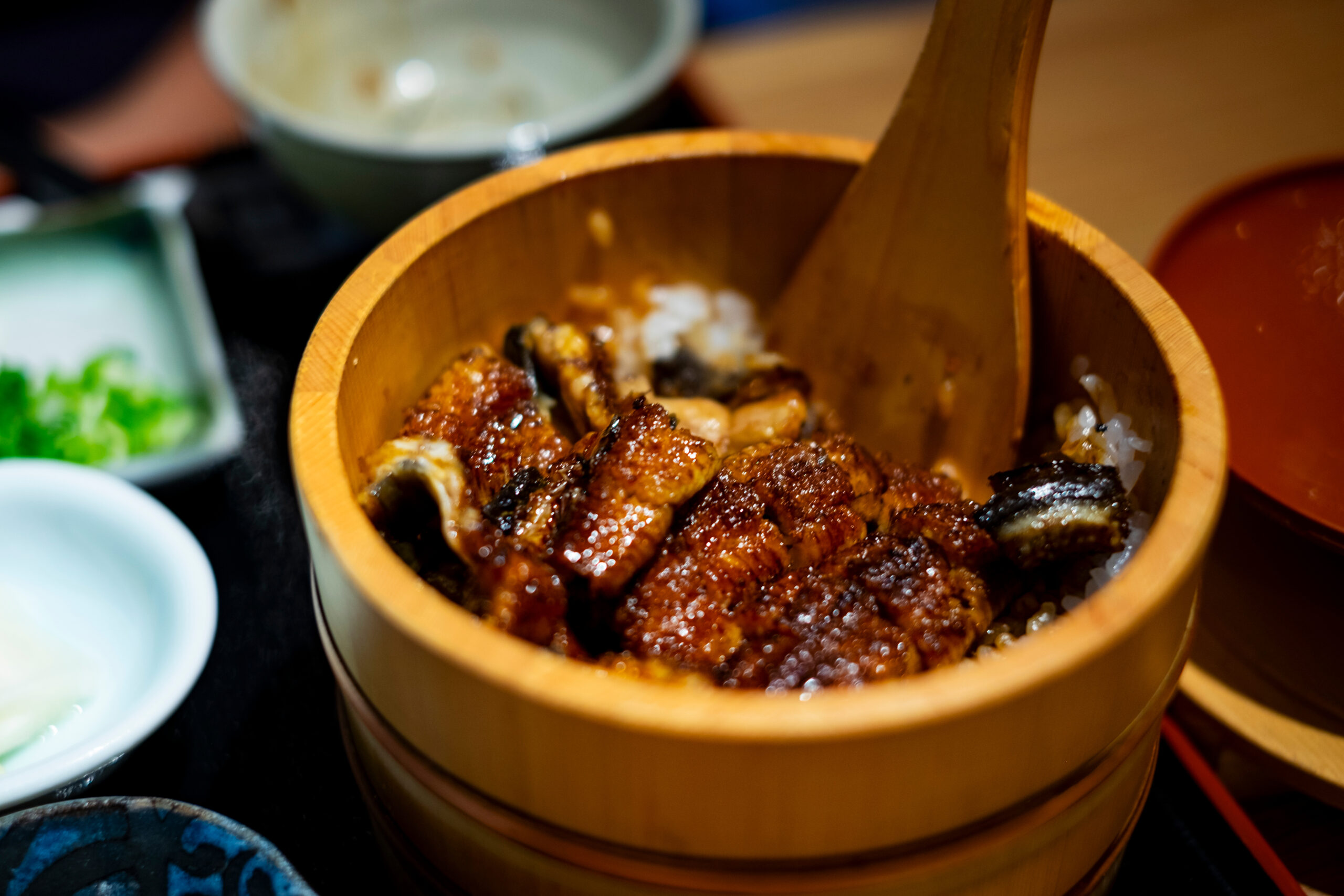 Wooden steamer basket filled with glossy glazed pork over white rice, with small side dishes nearby