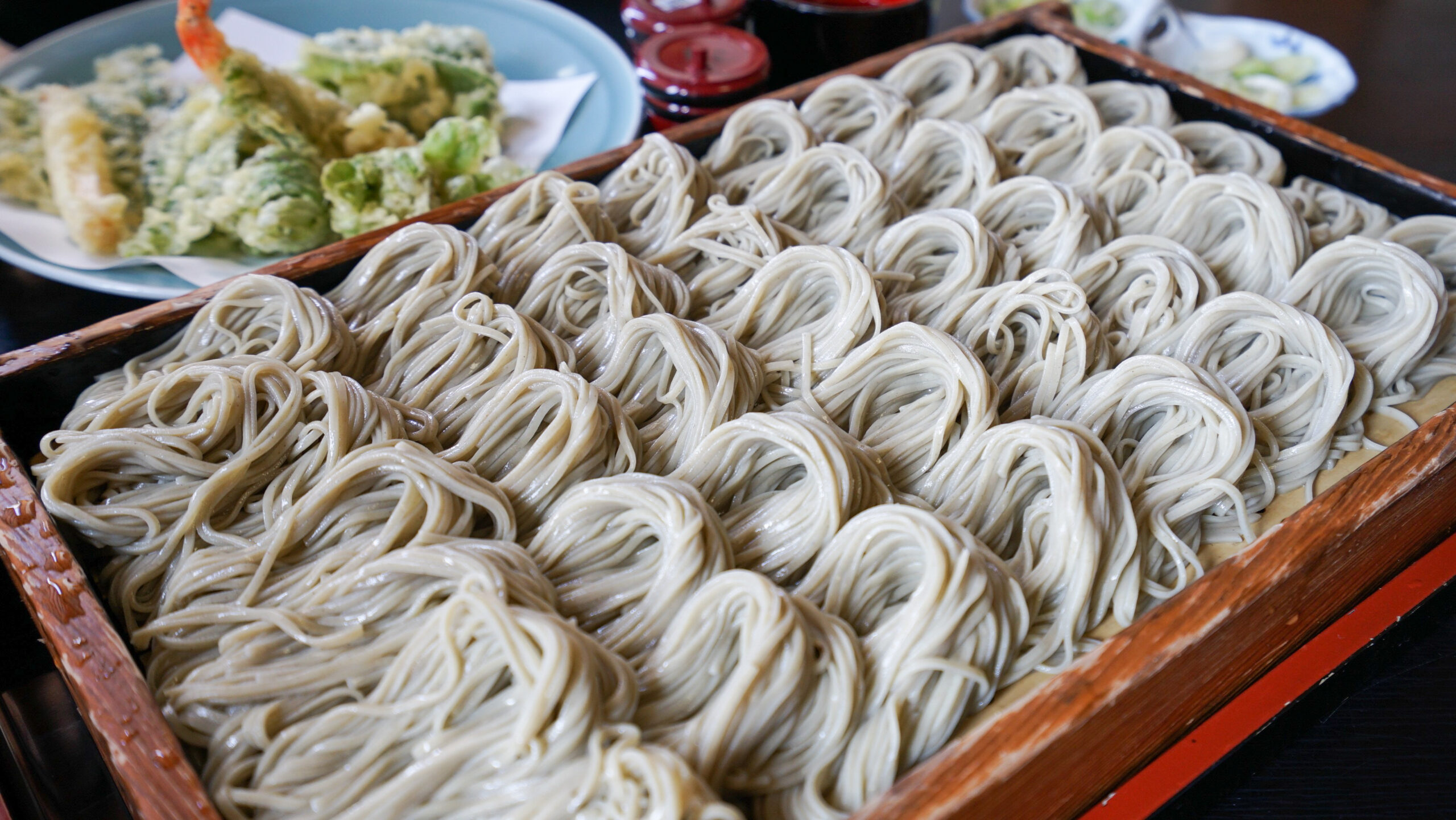 Wooden tray piled with pale soba noodles and a plate of tempura in the background, ready to serve