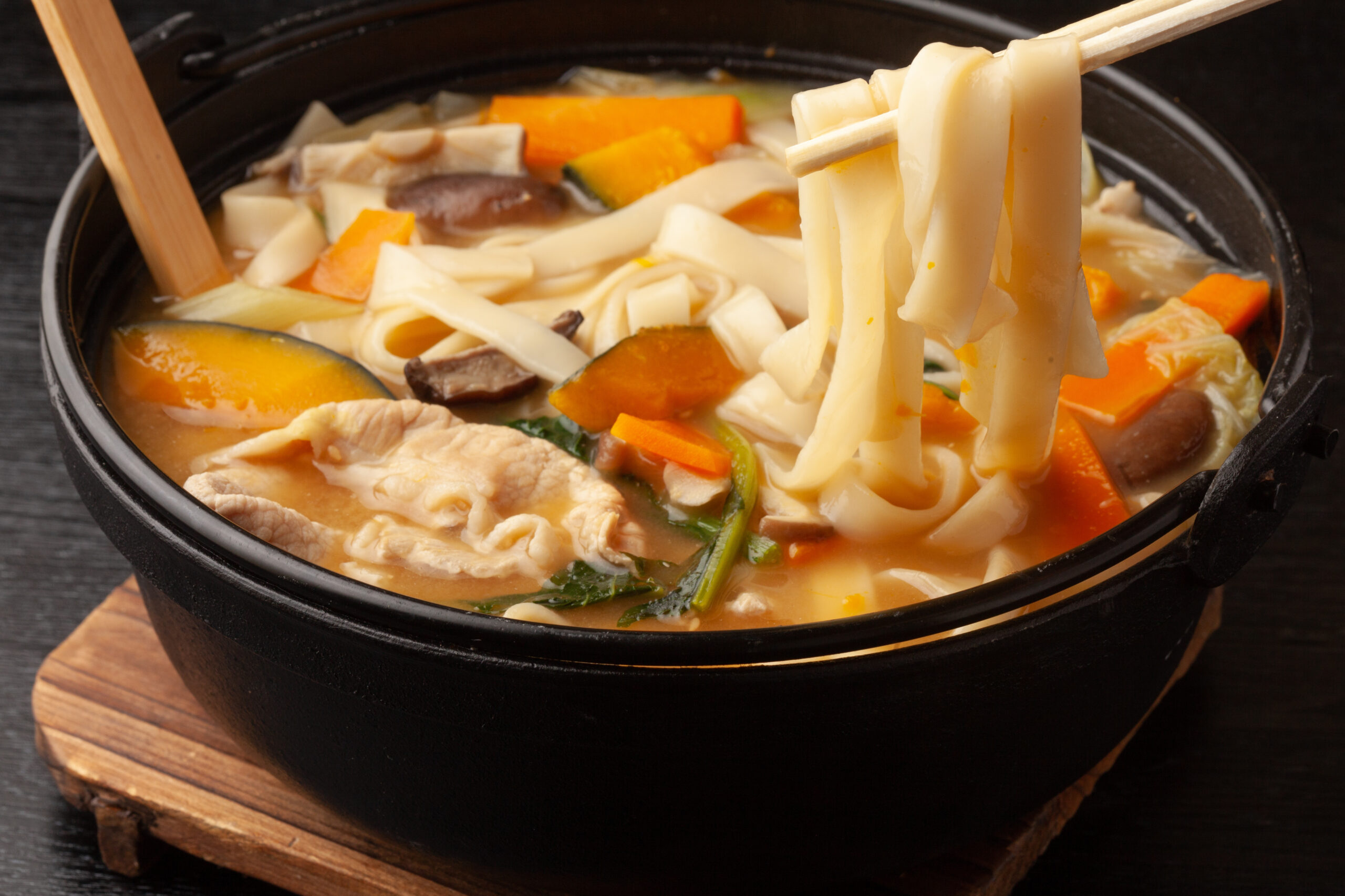 Noodle soup in a black pot with carrots, squash, mushrooms, and thin noodles, chopsticks lifting noodles.