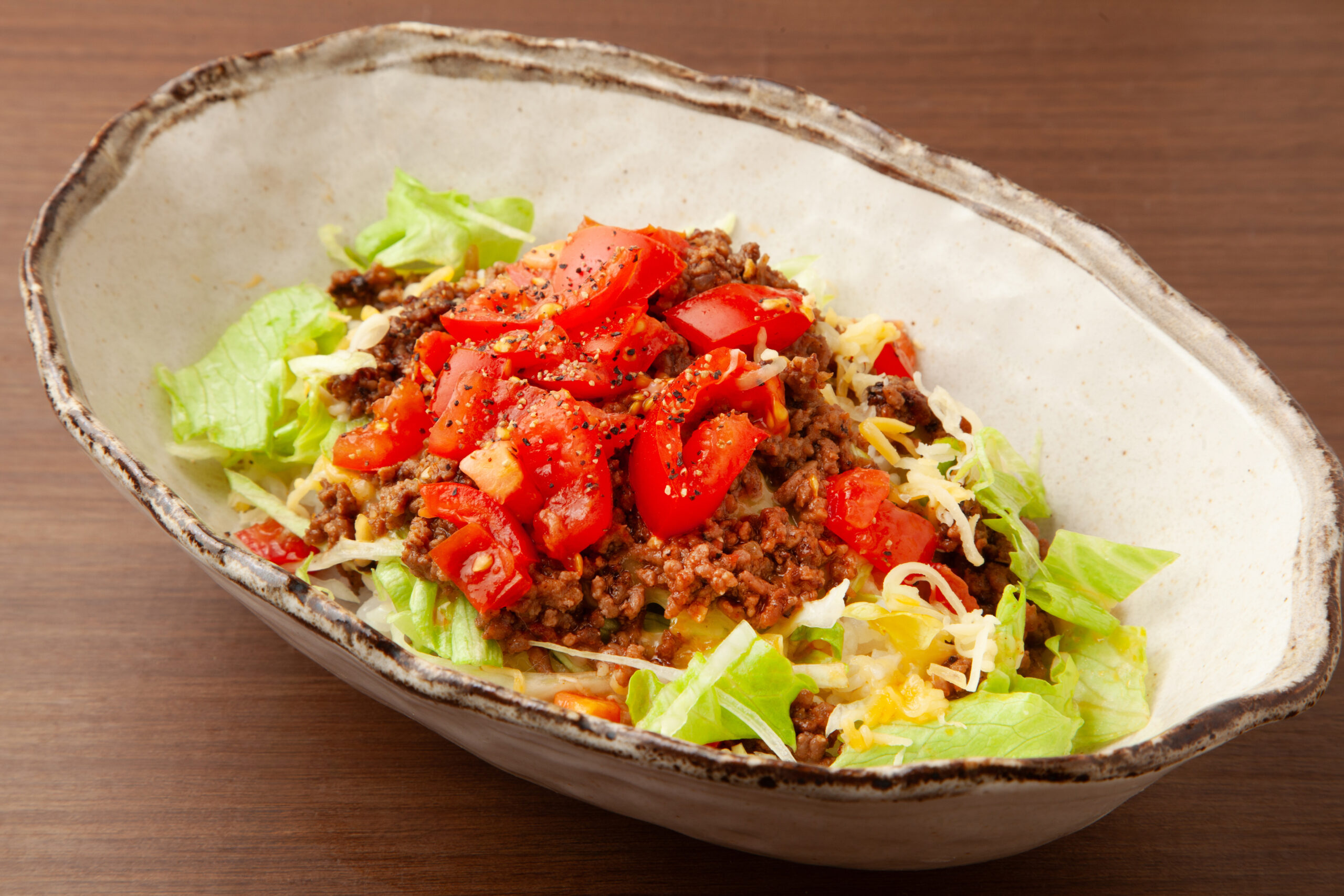Bowl of taco salad with ground beef, diced tomatoes, shredded cheese, and lettuce in a rustic ceramic bowl on a wooden table.