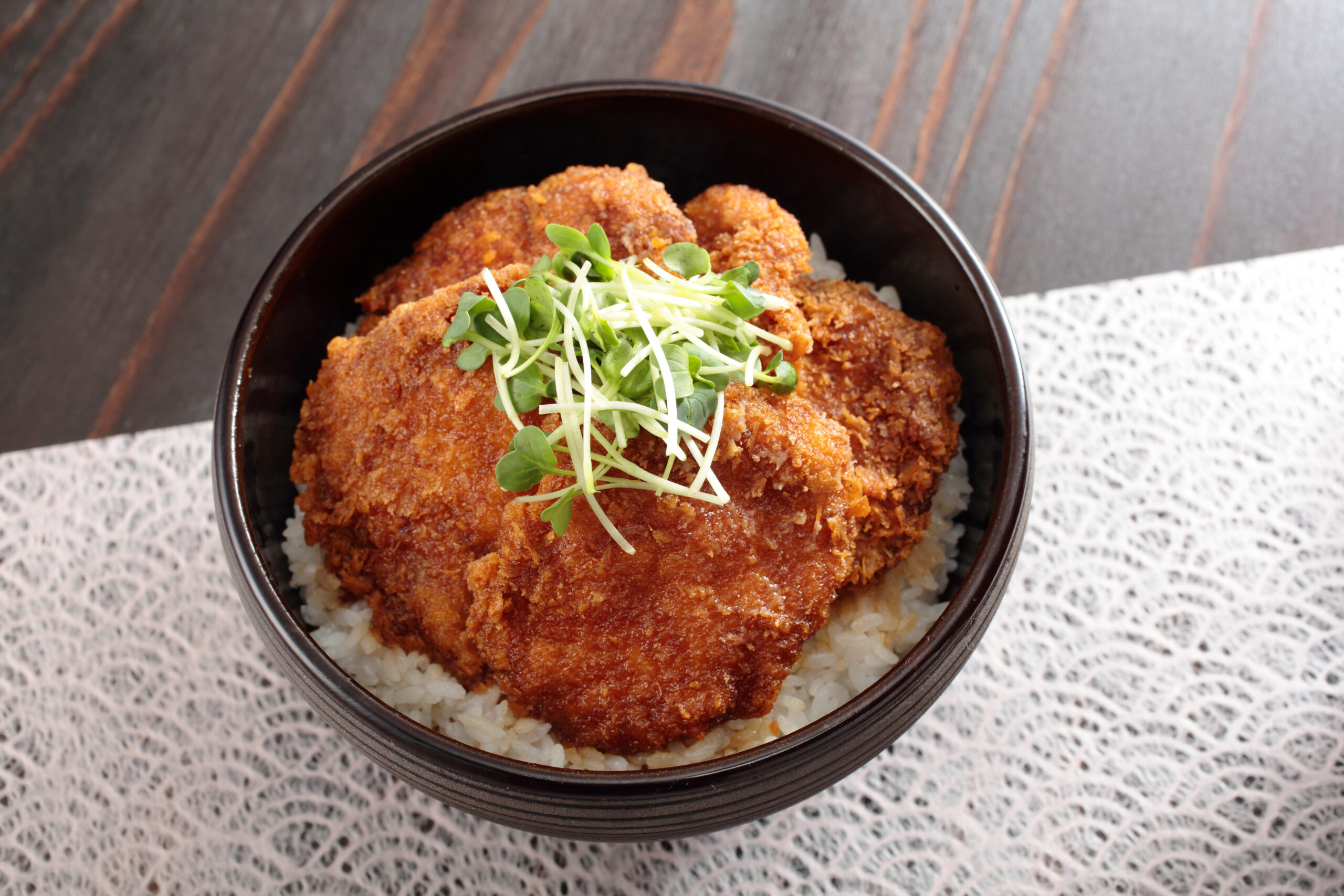 Crispy breaded fried meat on rice, garnished with microgreens in a dark bowl.