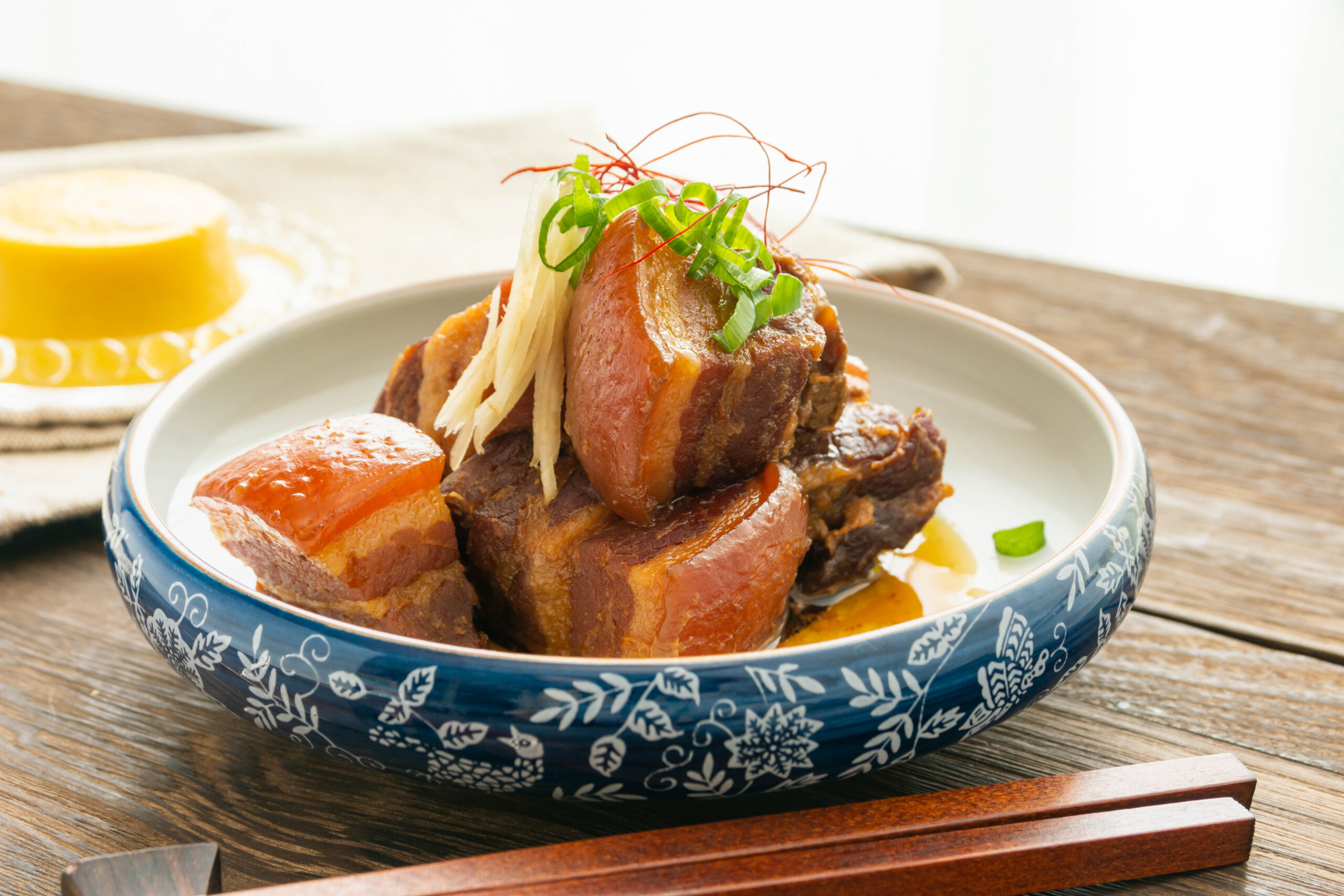 Braised pork with carrots and noodles in a blue patterned bowl, topped with green onions and red chili threads on a wooden table with chopsticks nearby.