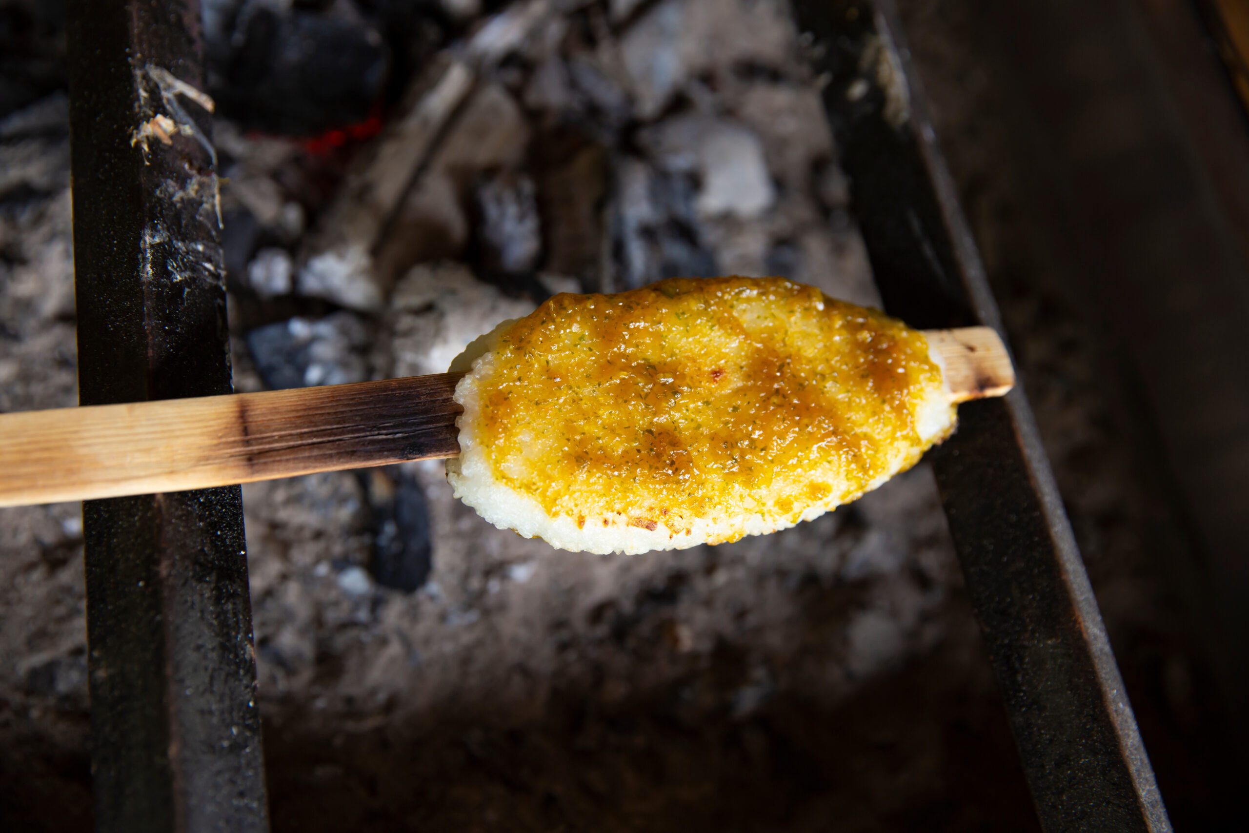 Grilled corn on the cob brushed with butter on a wooden skewer over charcoal grill bars.