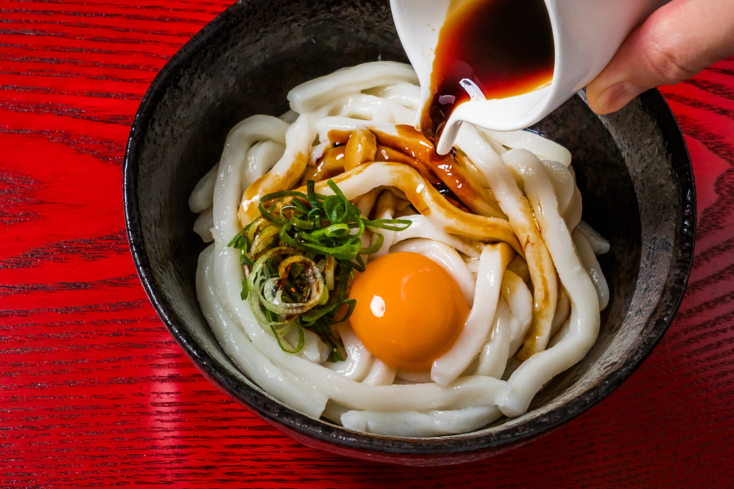 Thick udon noodles in a black bowl with green onions, an orange egg yolk, and sauce being poured over them by a hand on a red table.