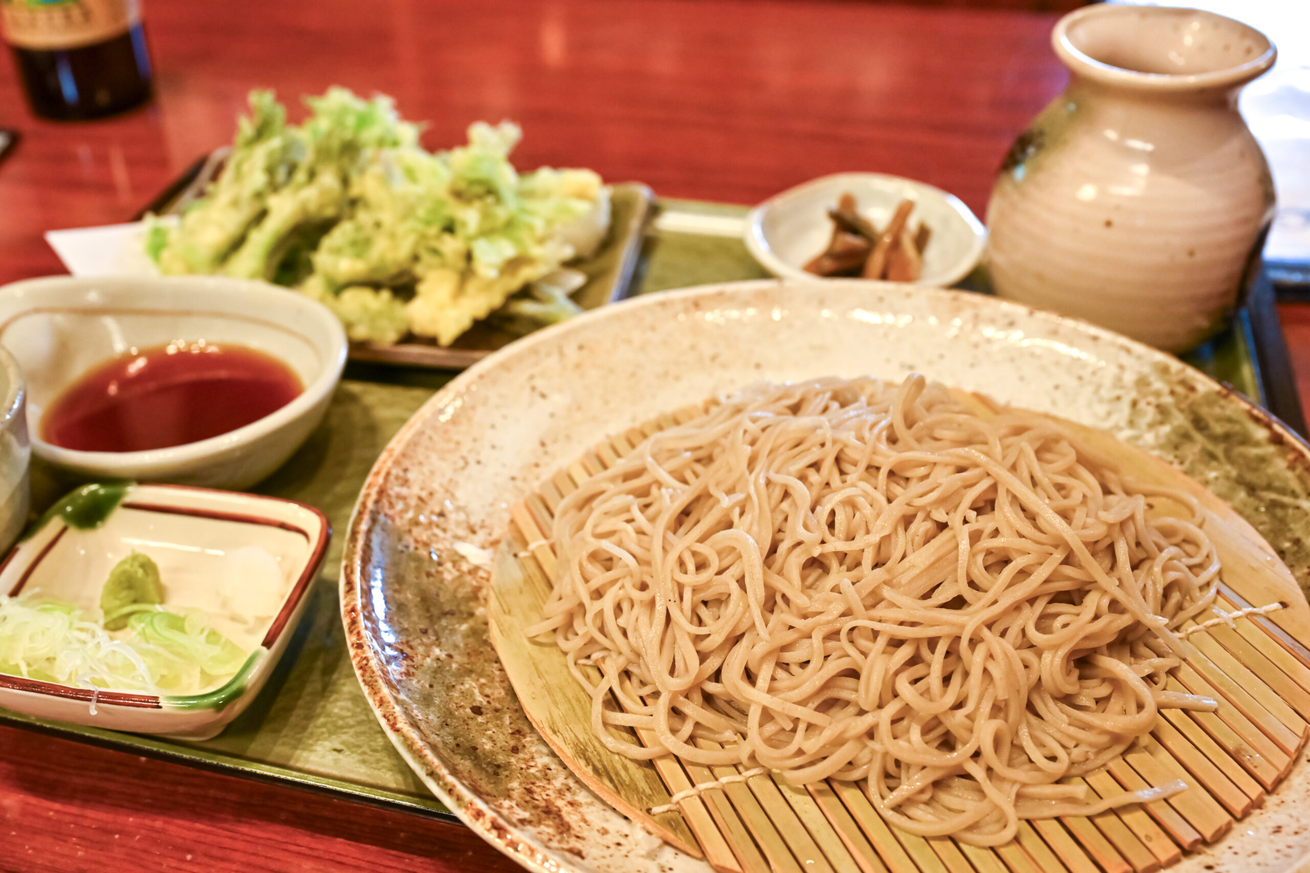 Soba noodles on a ceramic plate with a bamboo mat, dipping sauce in a small bowl, and tempura greens in the background.