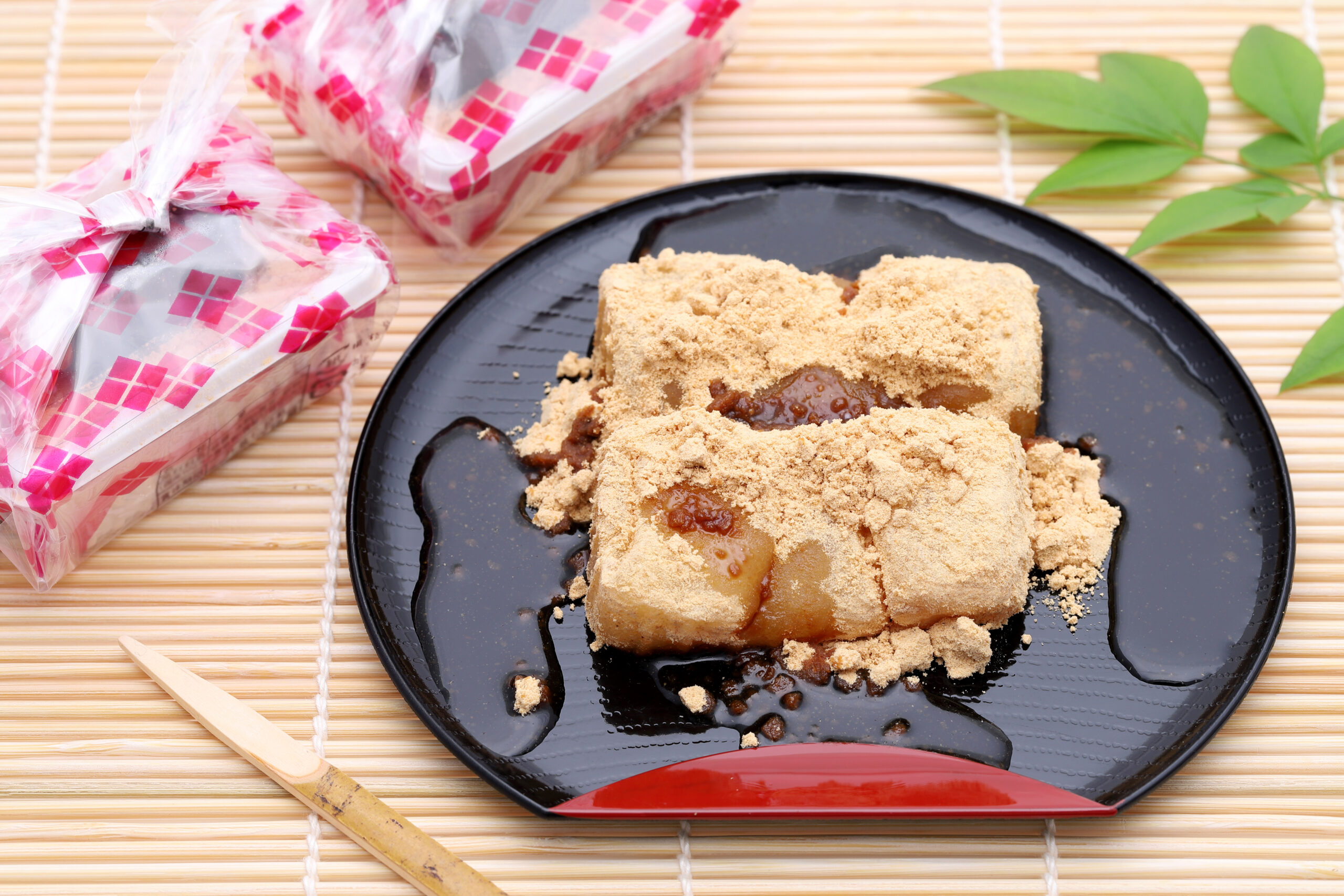 Mochi dessert dusted with kinako powder on a black plate, with pink-patterned gift boxes nearby on a bamboo mat surface.