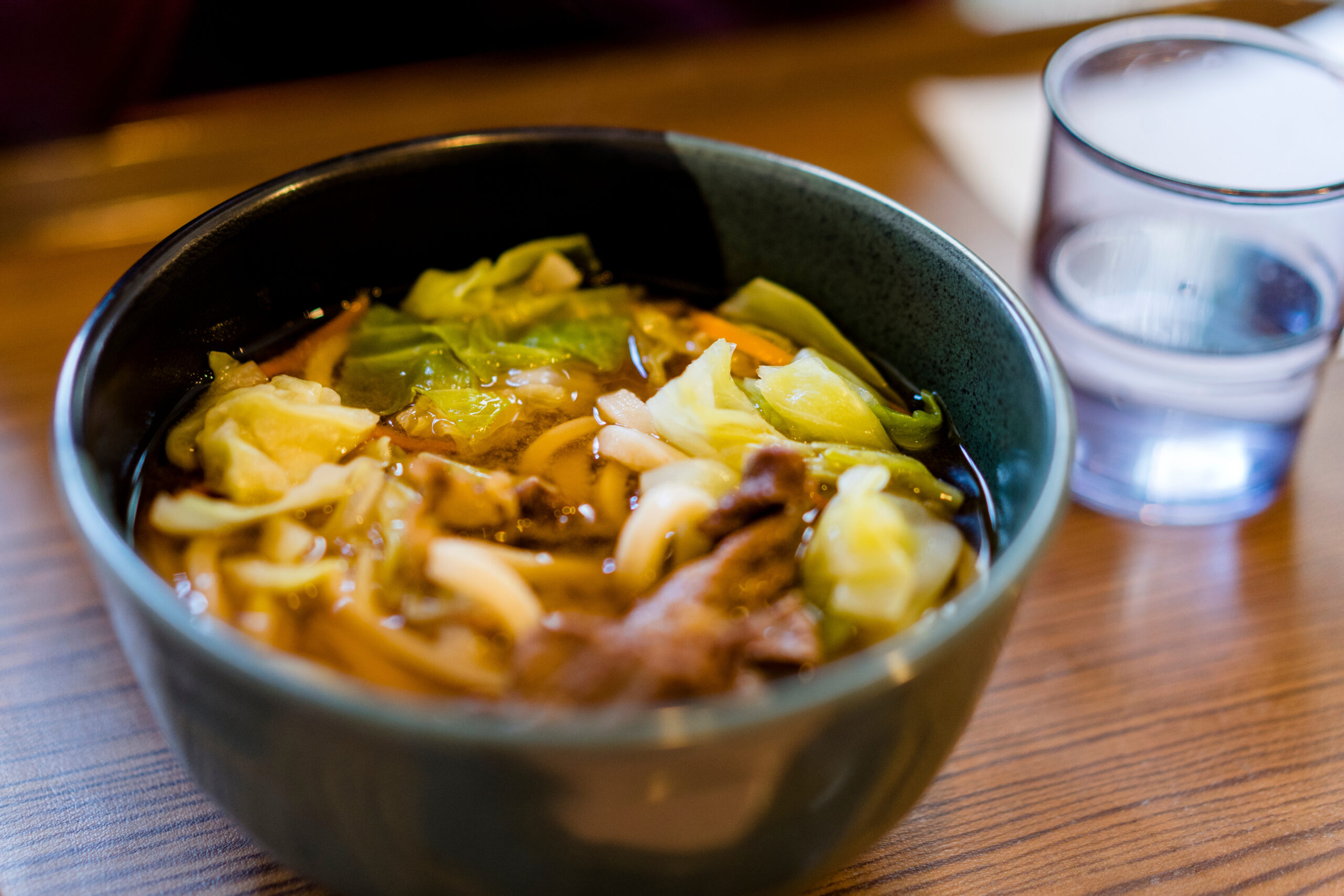 Bowl of vegetable soup with cabbage and meat chunks on a wooden table, with a glass of water to the right.