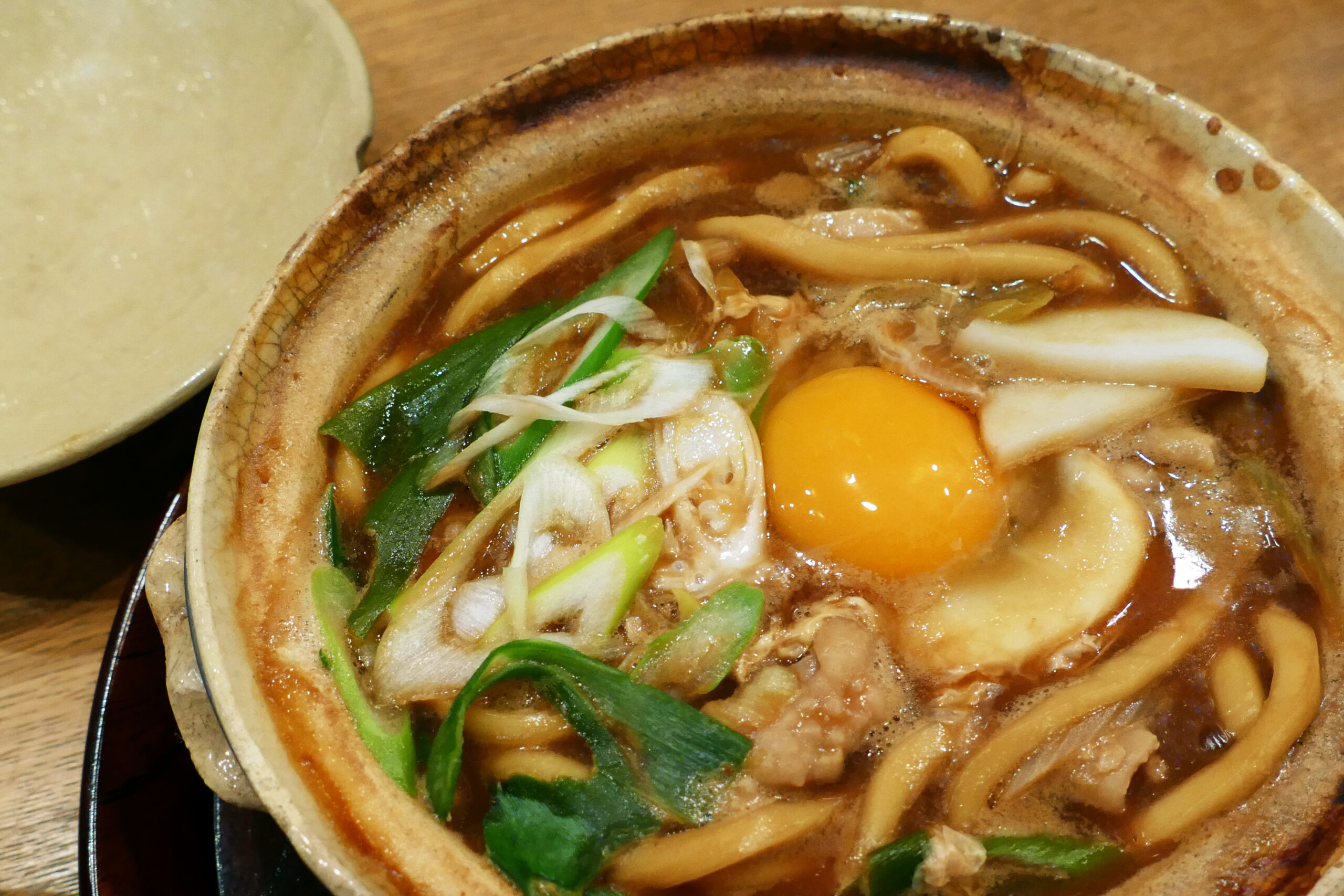 Close-up of a brown ceramic bowl of udon noodle soup with greens, onions, and a bright orange egg yolk top.