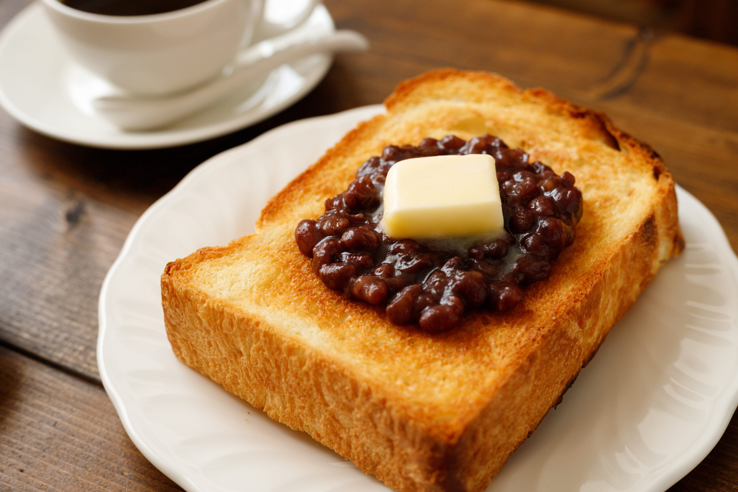Toast with butter on top of sweet red bean paste, served on a white plate with a coffee cup in the background.
