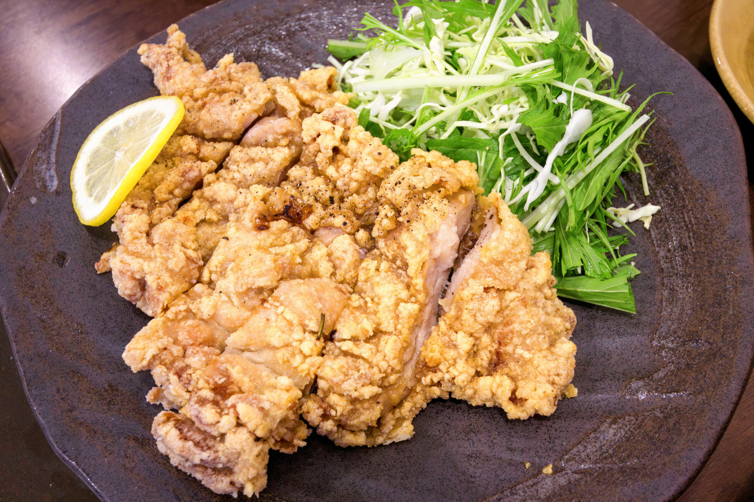 Breaded fried chicken pieces with a lemon half on a dark plate, accompanied by a pile of shredded greens on the side.