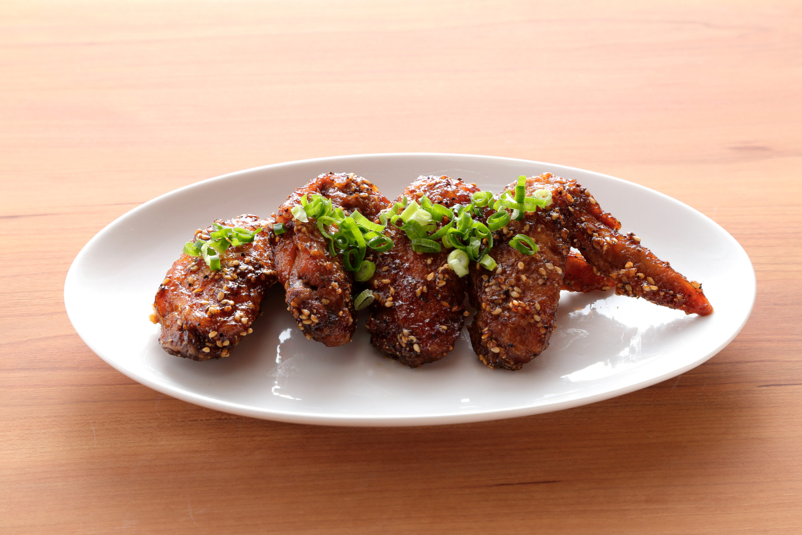 Plate of glazed chicken wings sprinkled with sesame seeds and chopped green onions on a wooden table.