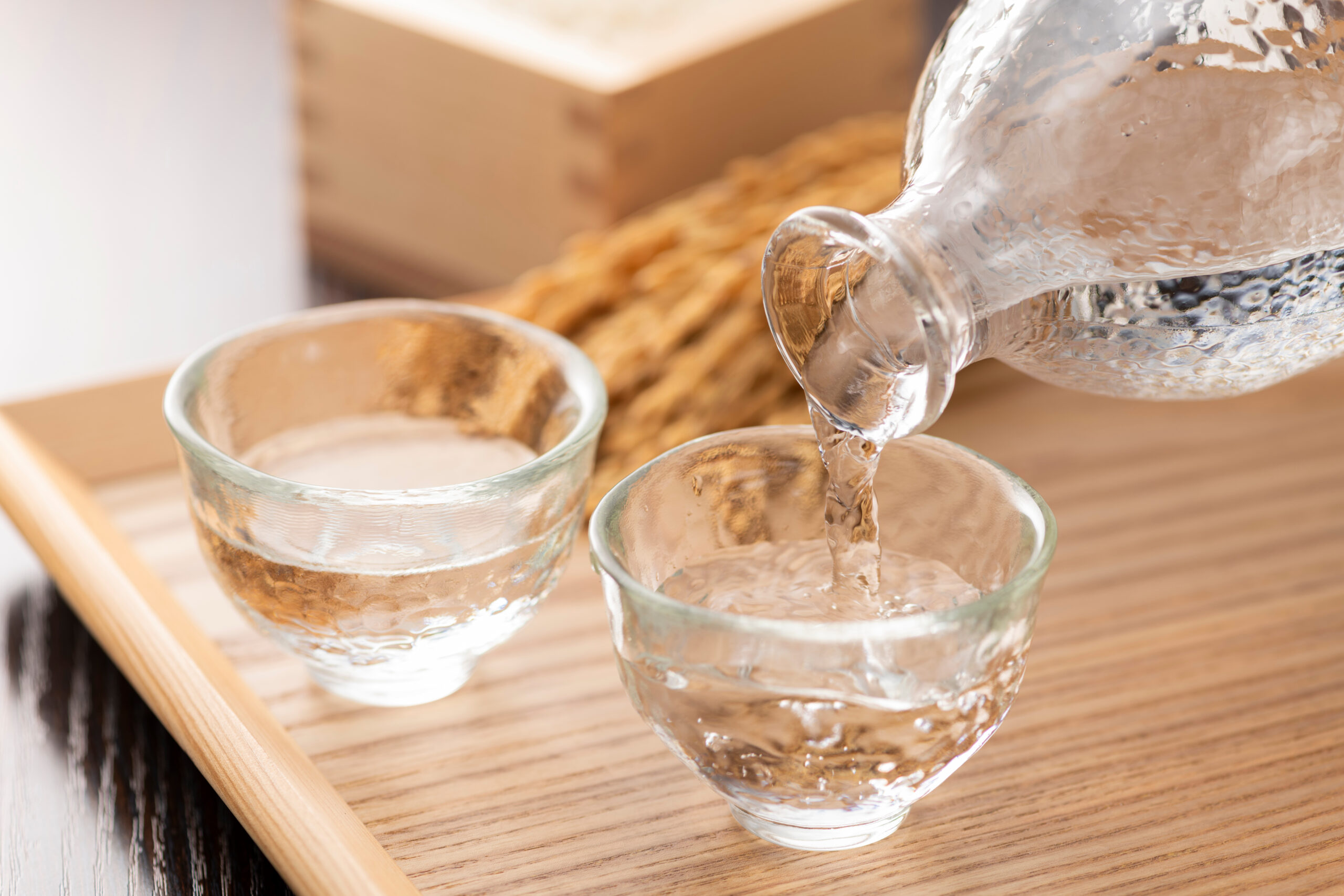 Pouring clear liquid into small glass sake cups on a wooden tray.