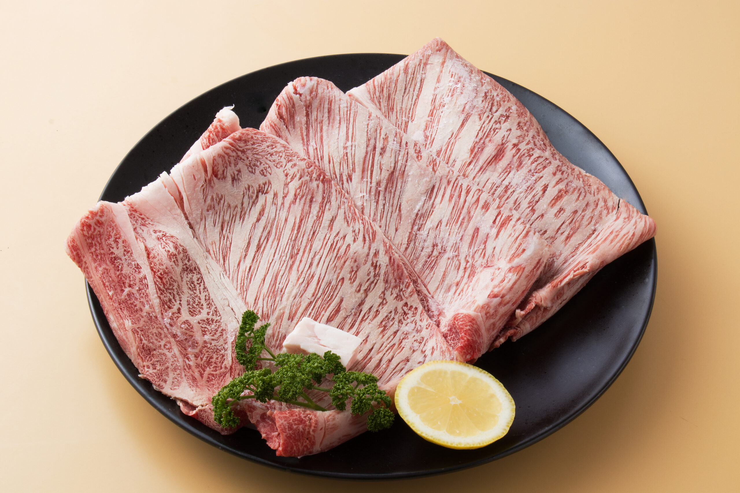 Thinly sliced marbled beef on a black plate with lemon wedges and parsley sprigs.