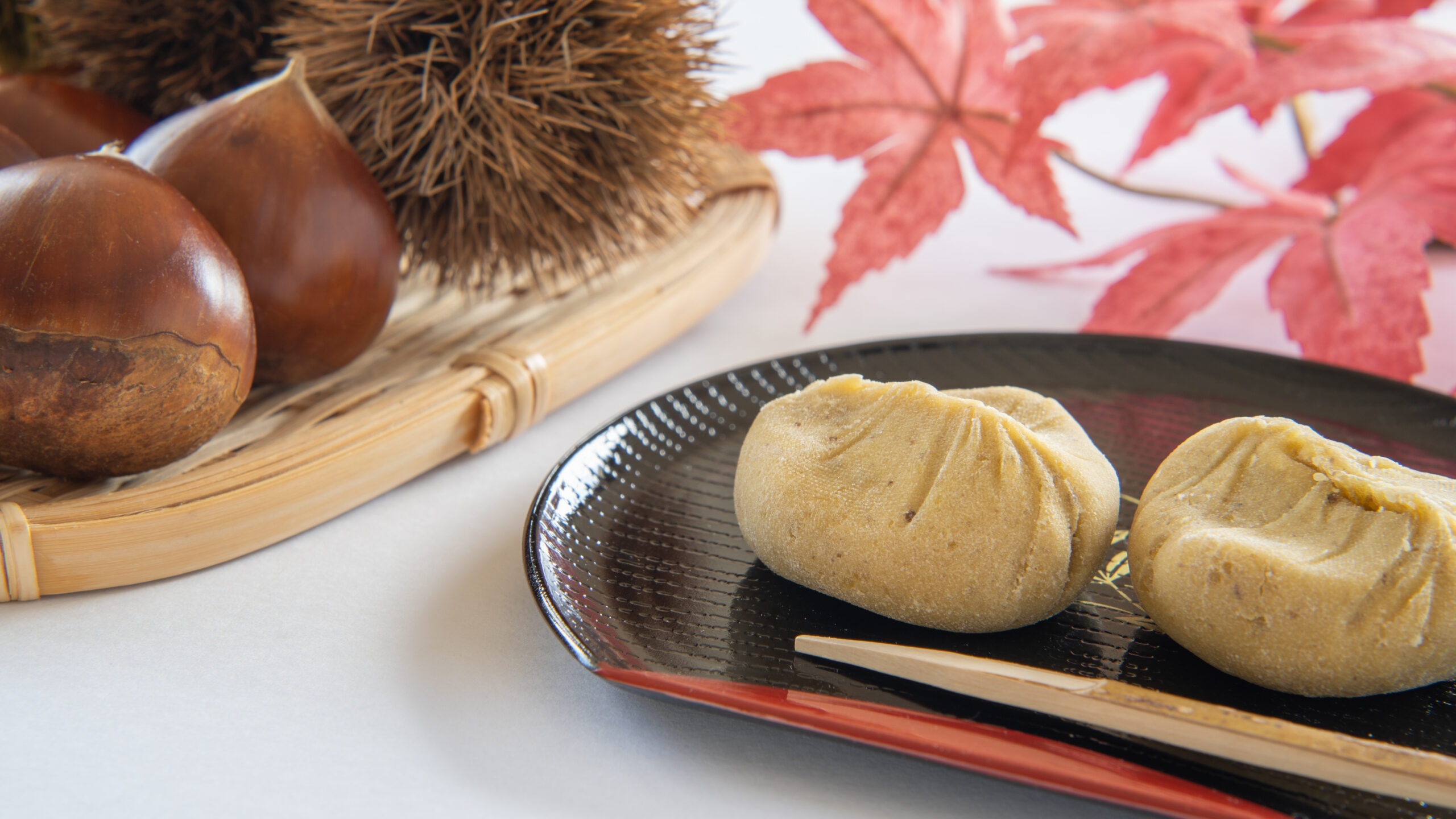Autumn still life with chestnuts in a woven basket, a black plate holding round dumplings, chopsticks, and red maple leaves in the background.