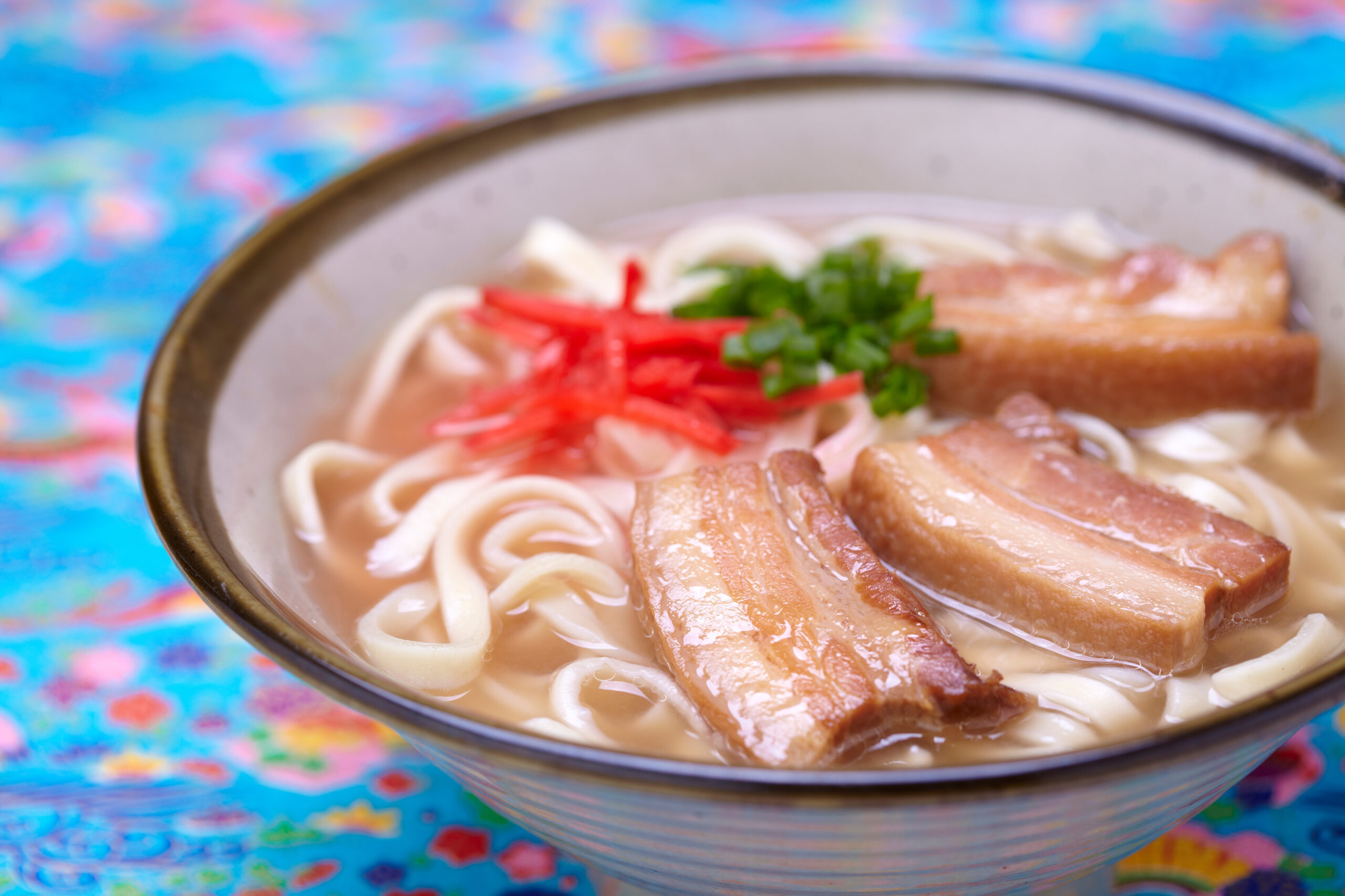 Bowl of ramen with sliced pork, noodles, green onions, and red pickled ginger in broth on a colorful table.