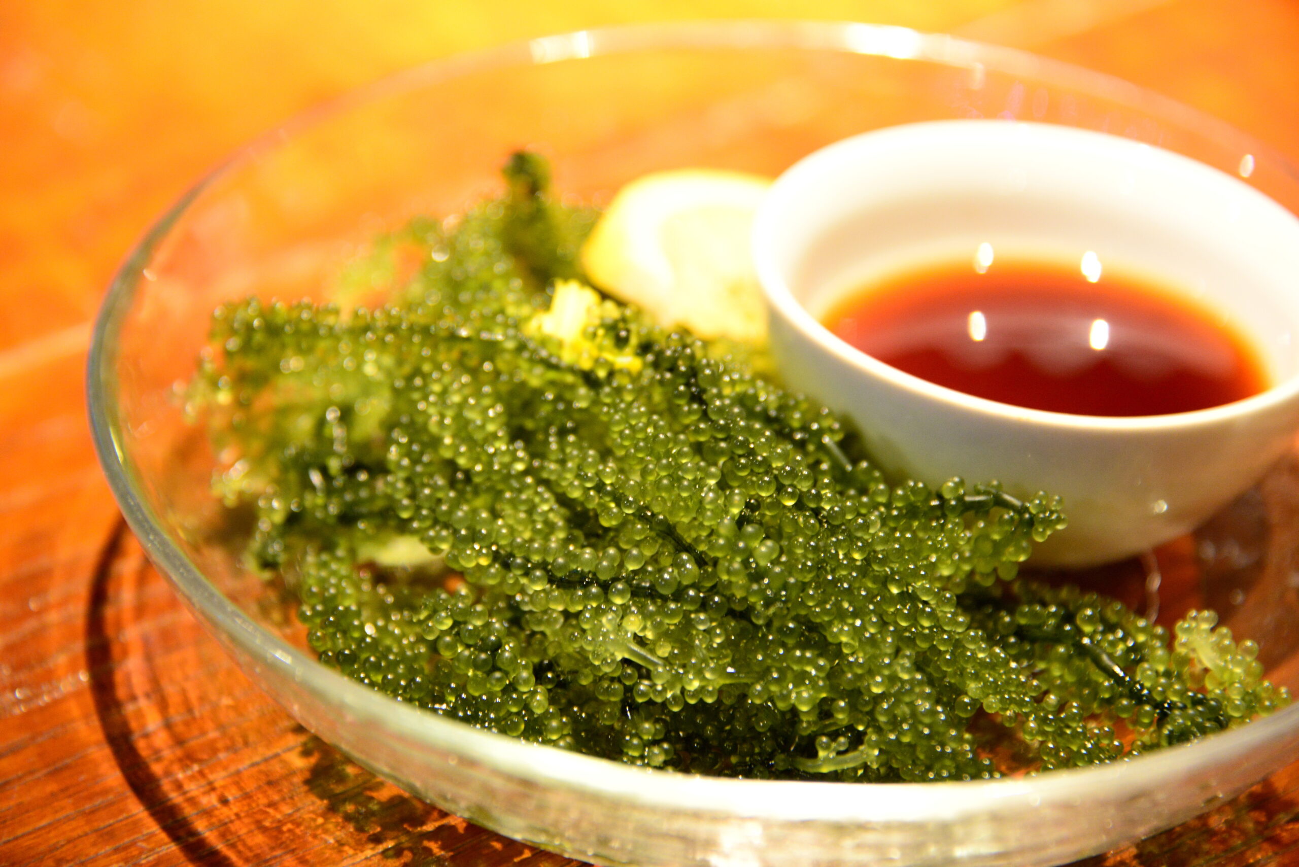 Bowl of green sea grapes (green caviar) with a small white cup of red dipping sauce on a wooden table.