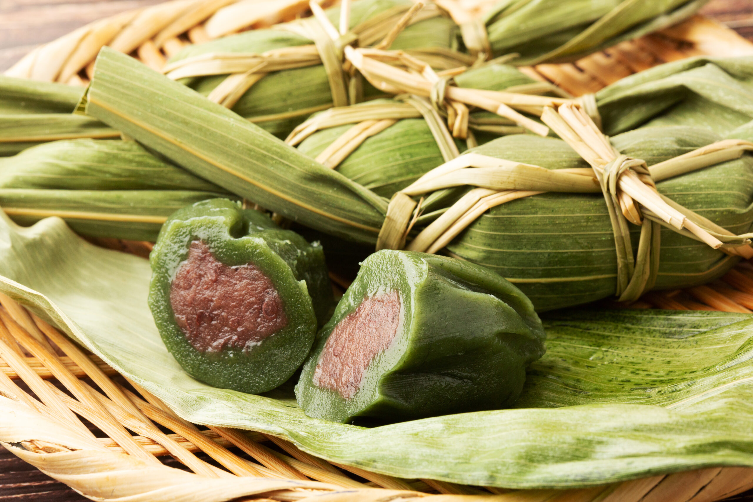 Bamboo leaf–wrapped glutinous rice dumplings tied with straw, arranged in a basket with one cut open to show a reddish filling inside the green leaf wrapper.