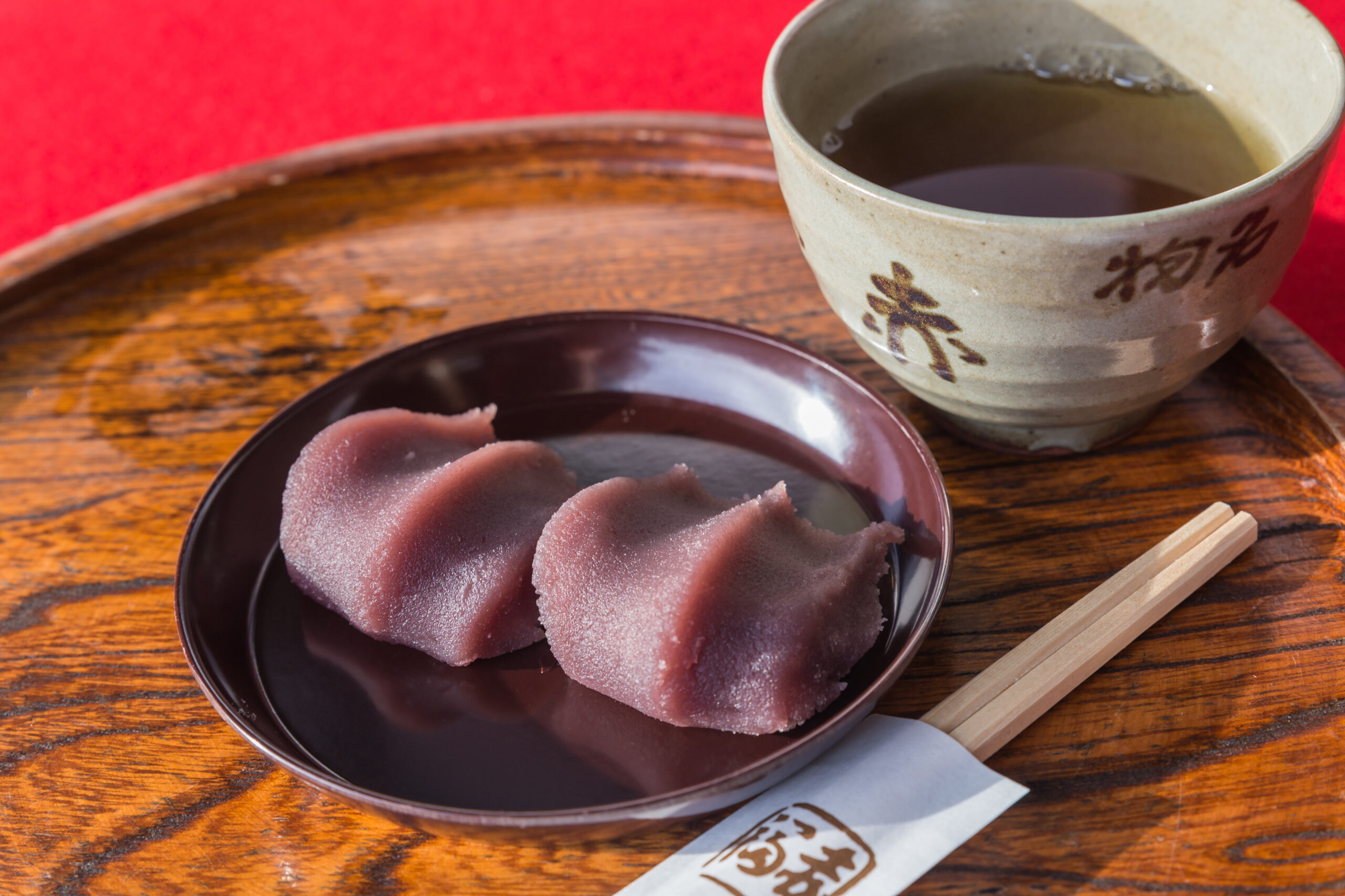 Pink mochi pieces on a dark plate, with a ceramic teacup and wooden tray on a red backdrop (Japanese dessert scene).