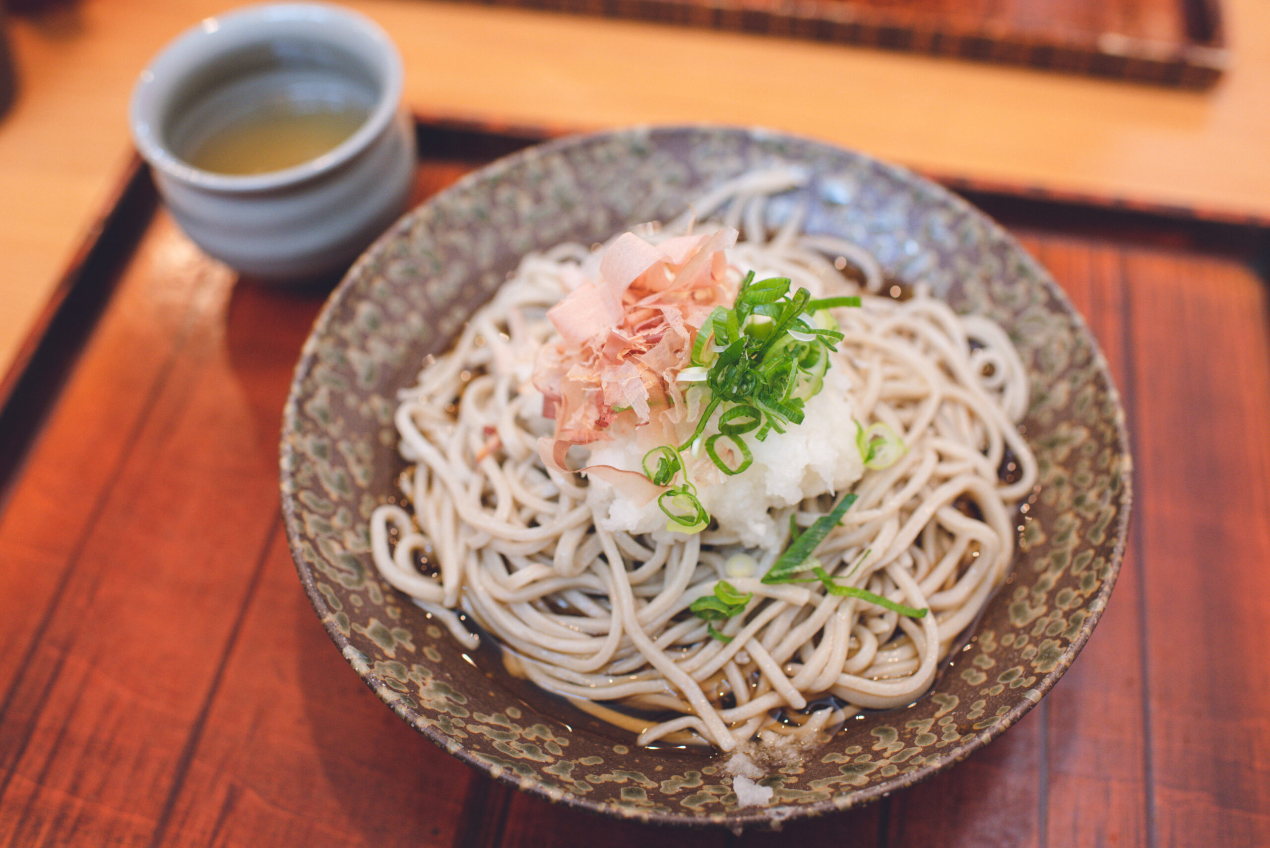 Bowl of soba noodles topped with green onions and bonito flakes, served with a small cup on the side.