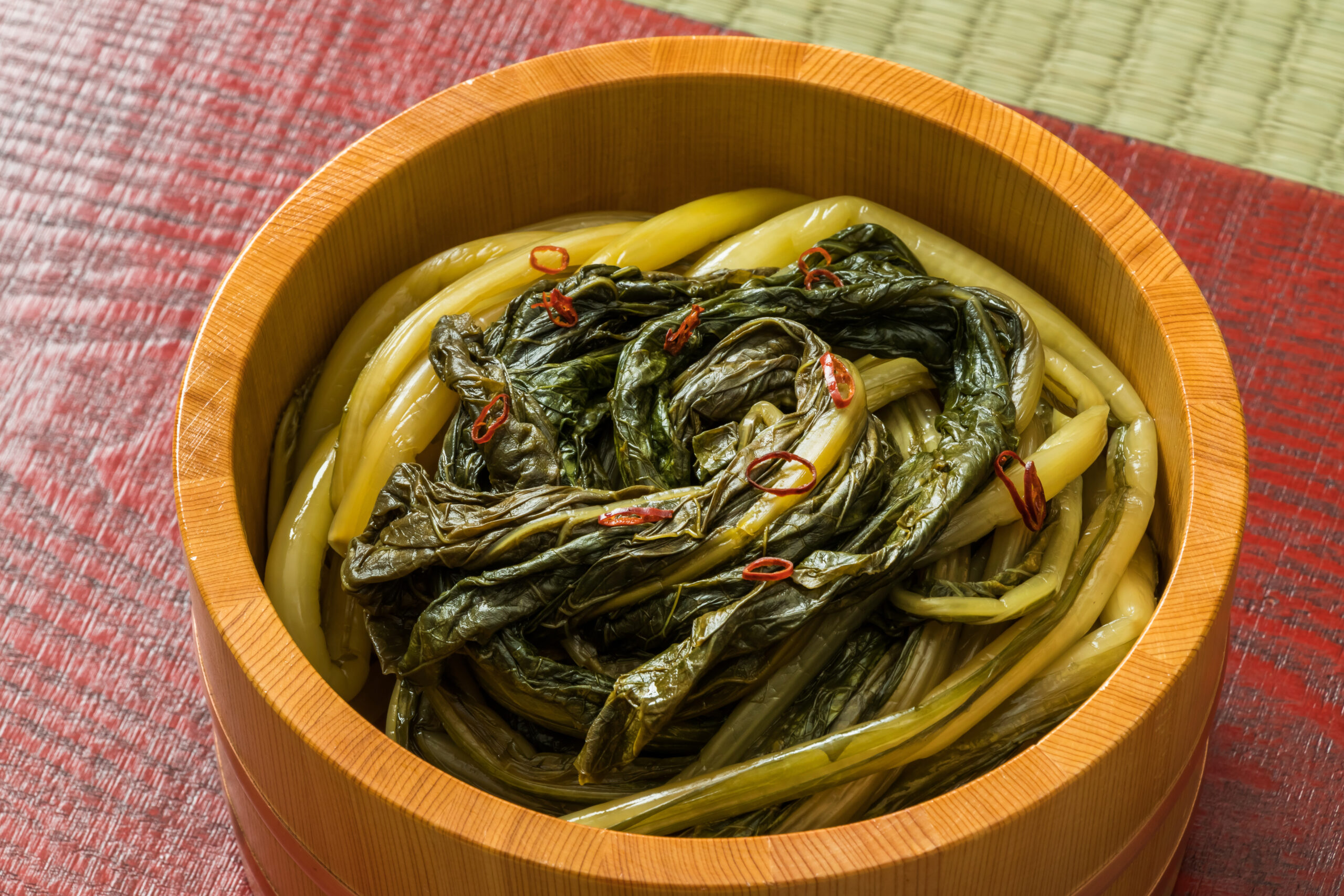 Wooden tub filled with pickled mustard greens and pale stalks, garnished with red chili rings.