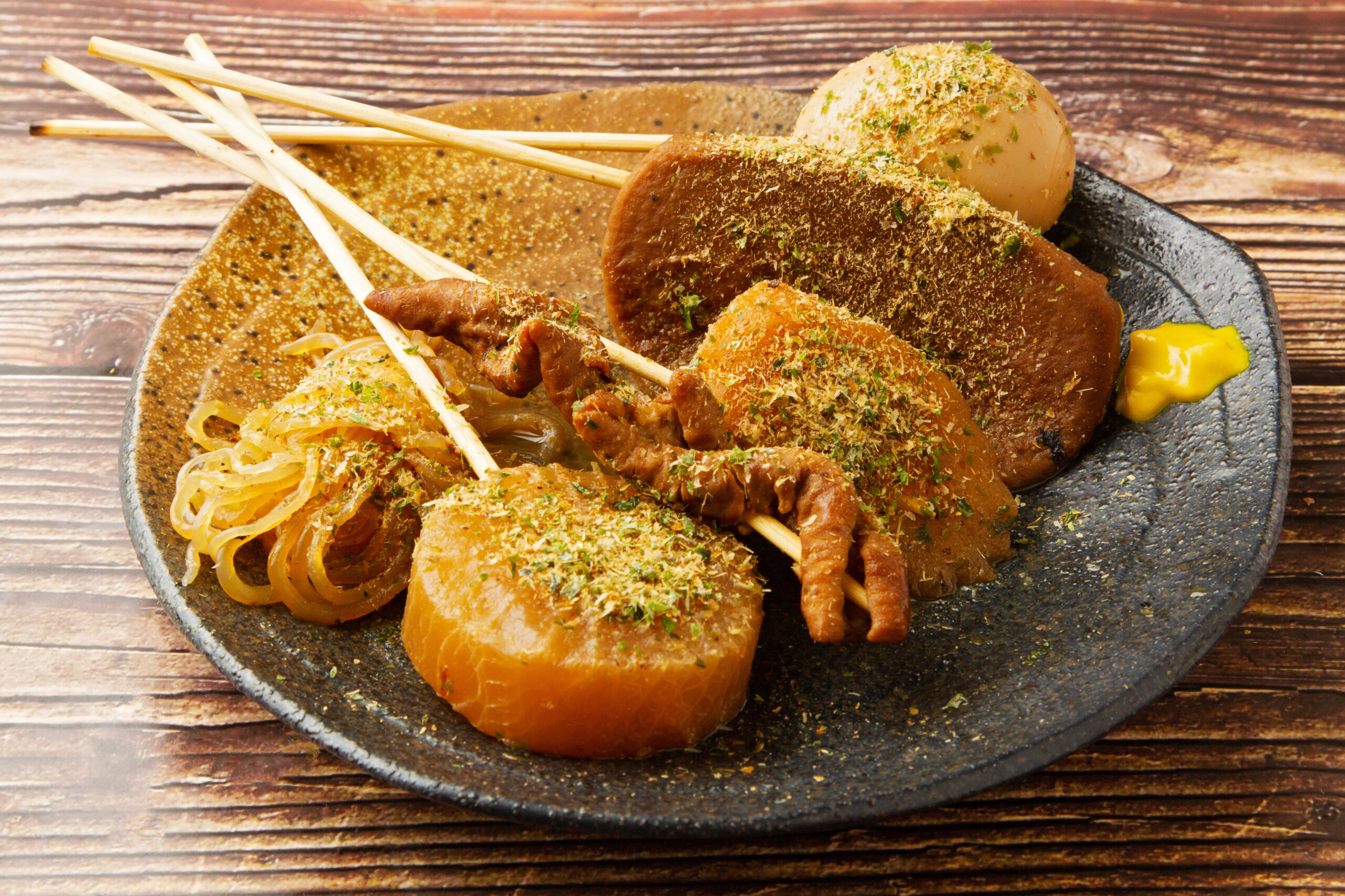 Skewered grilled meat with herbs on a plate beside noodles and a bread roll on a rustic wooden table.