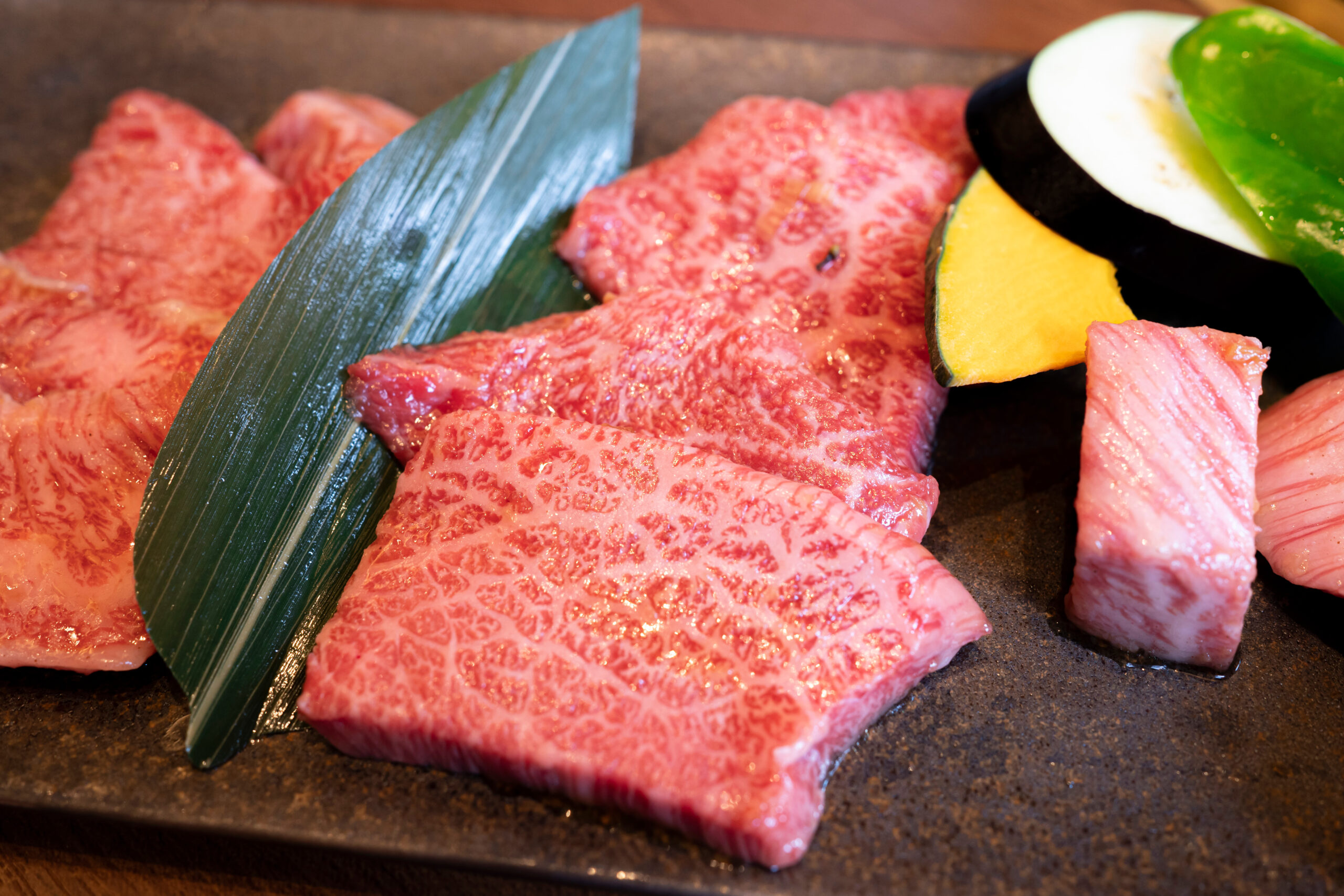 Slices of heavily marbled raw beef on a grill pan, with a blue-green leaf and a slice of yellow squash nearby as garnish.
