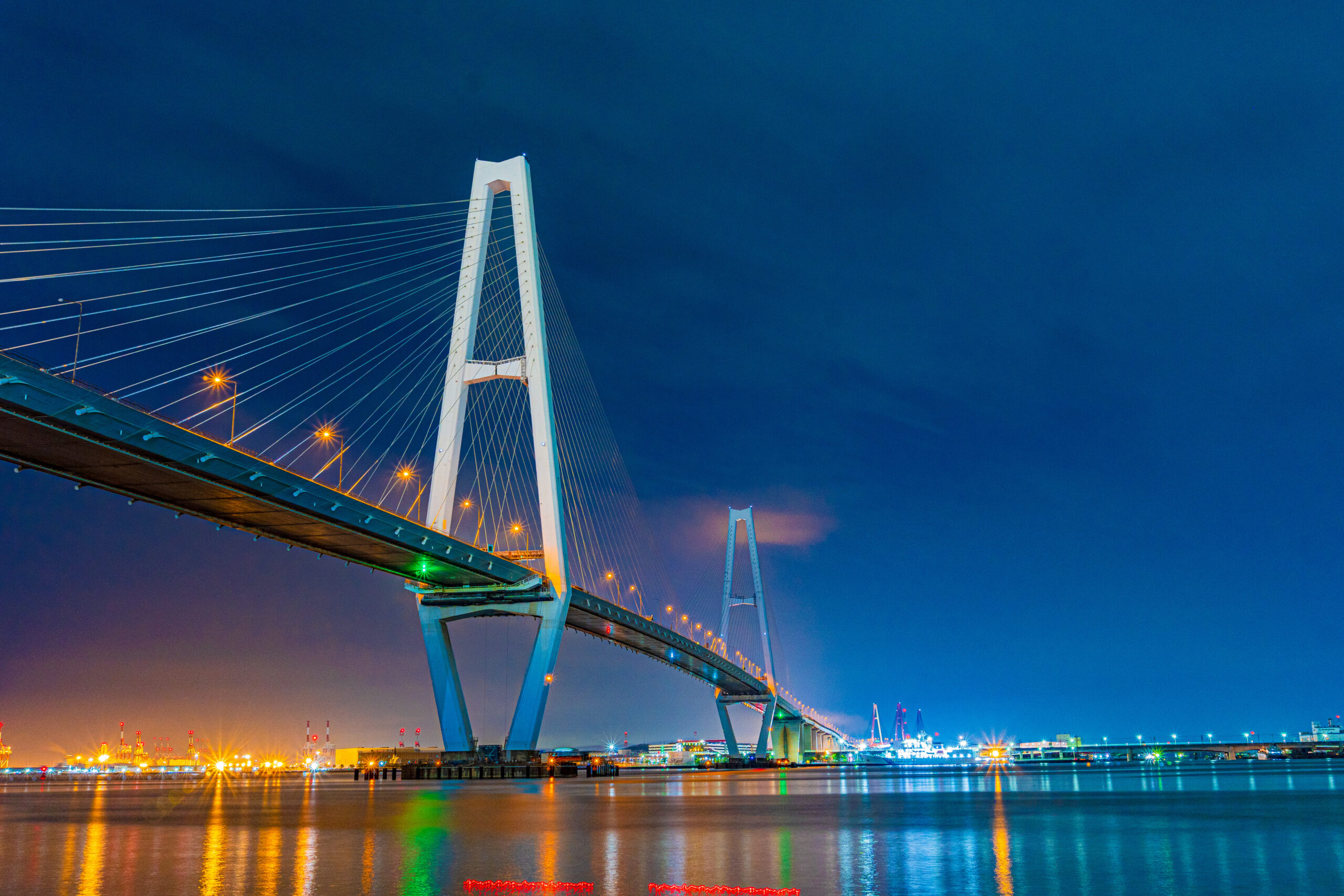 Cable-stayed bridge at night with blue sky, illuminated pylons and numerous suspension cables reflecting on calm water
