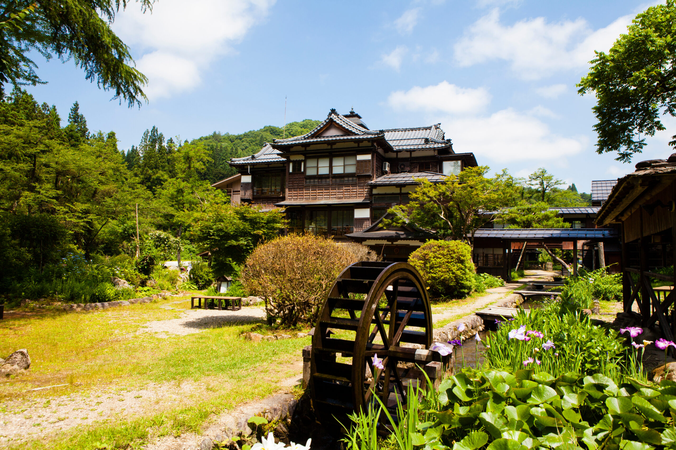 Traditional wooden Japanese house with a tiled roof beside a large water wheel in a lush garden.