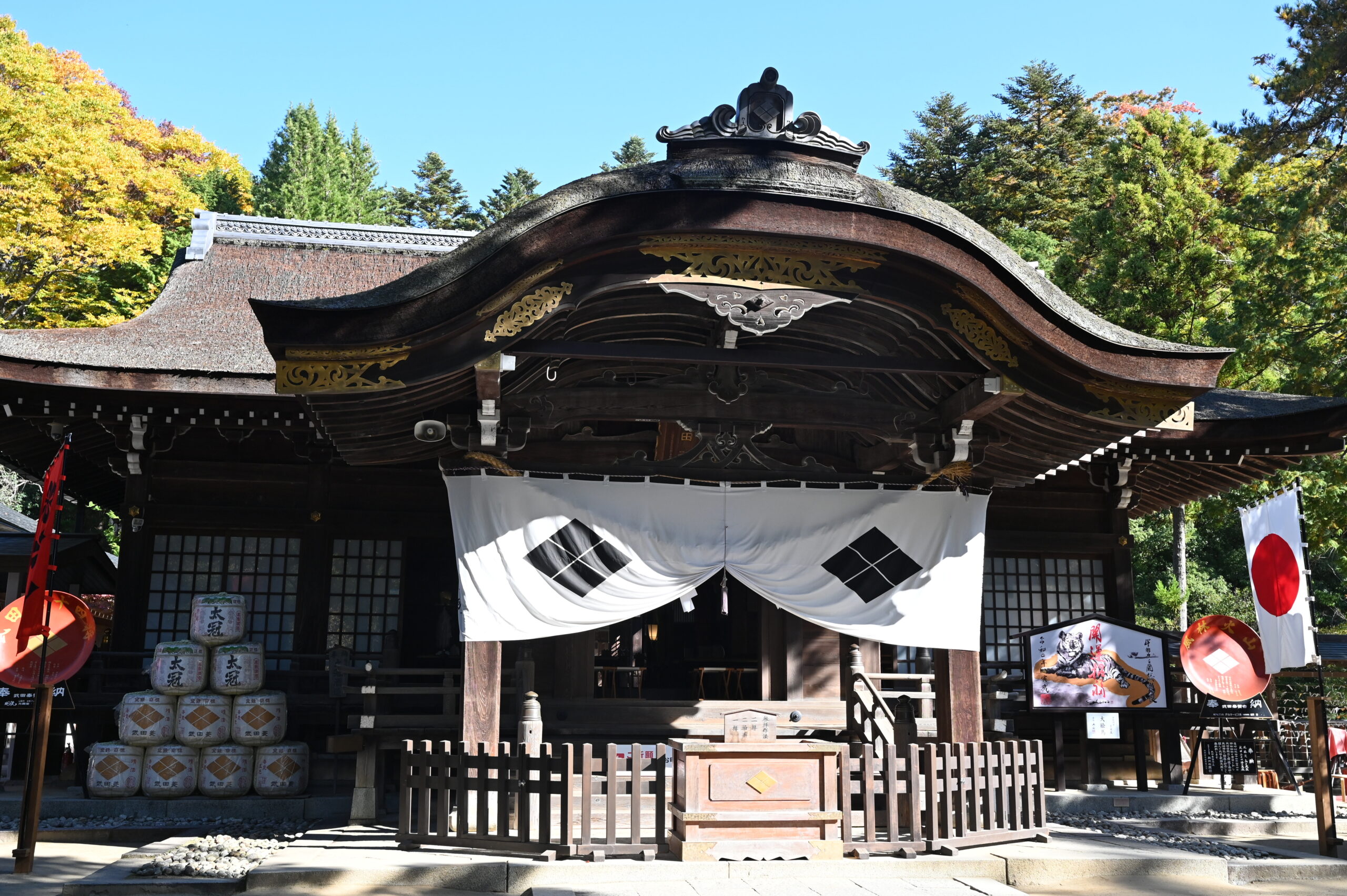 Traditional Japanese shrine with a curved thatched roof, white fabric banners, and ornate gold trim, set among autumn trees.