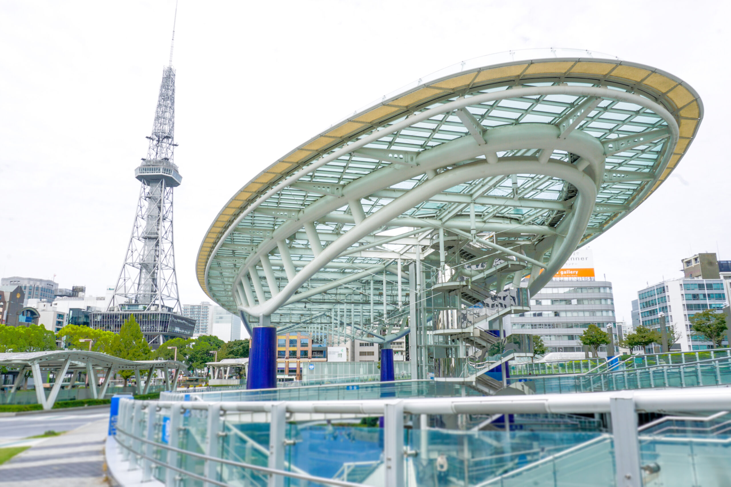 Curved glass and metal canopy over a walkway in a modern urban plaza with a tall lattice tower in the background.