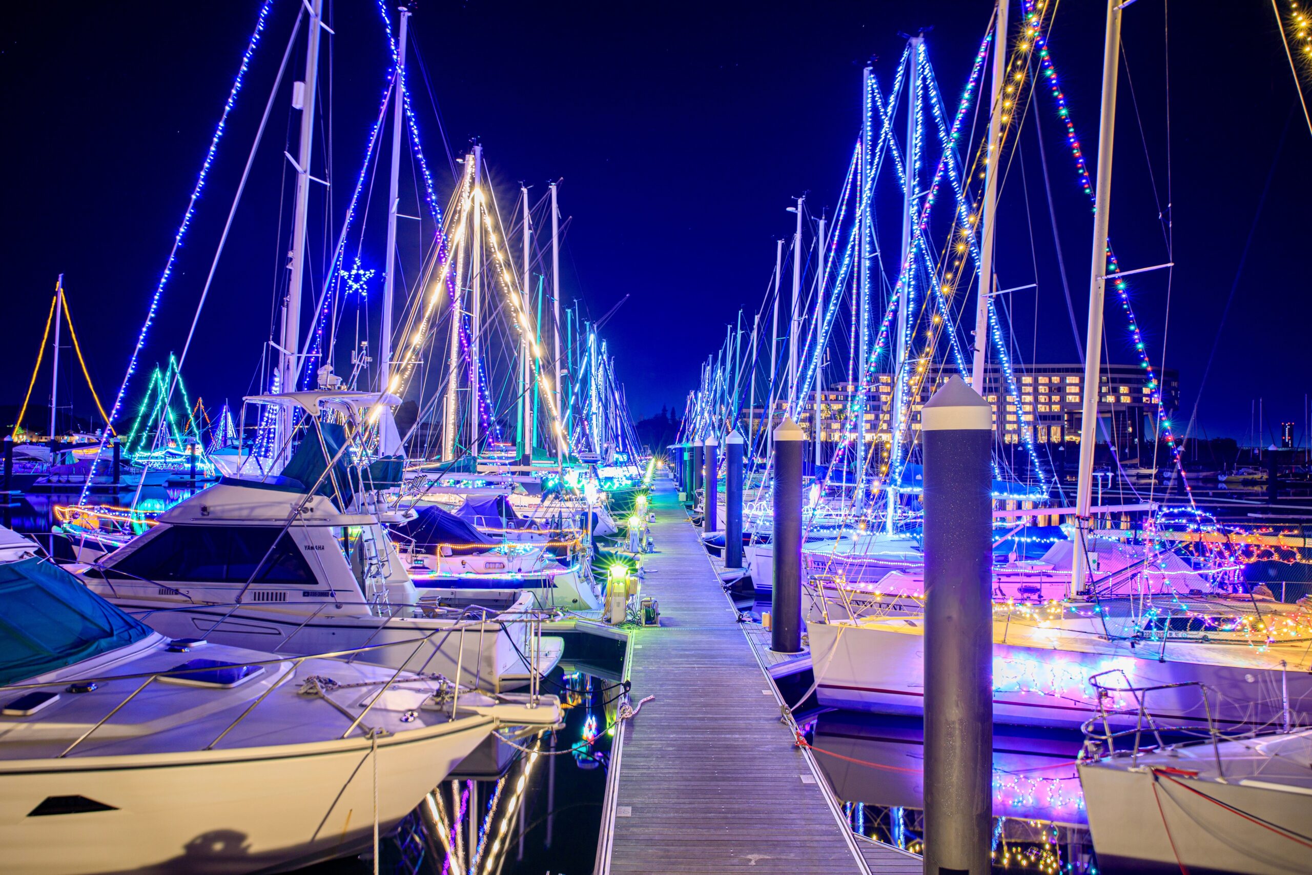 Marina at night with sailboats docked along a pier, rigging wrapped in blue and multicolored lights reflecting on the water
