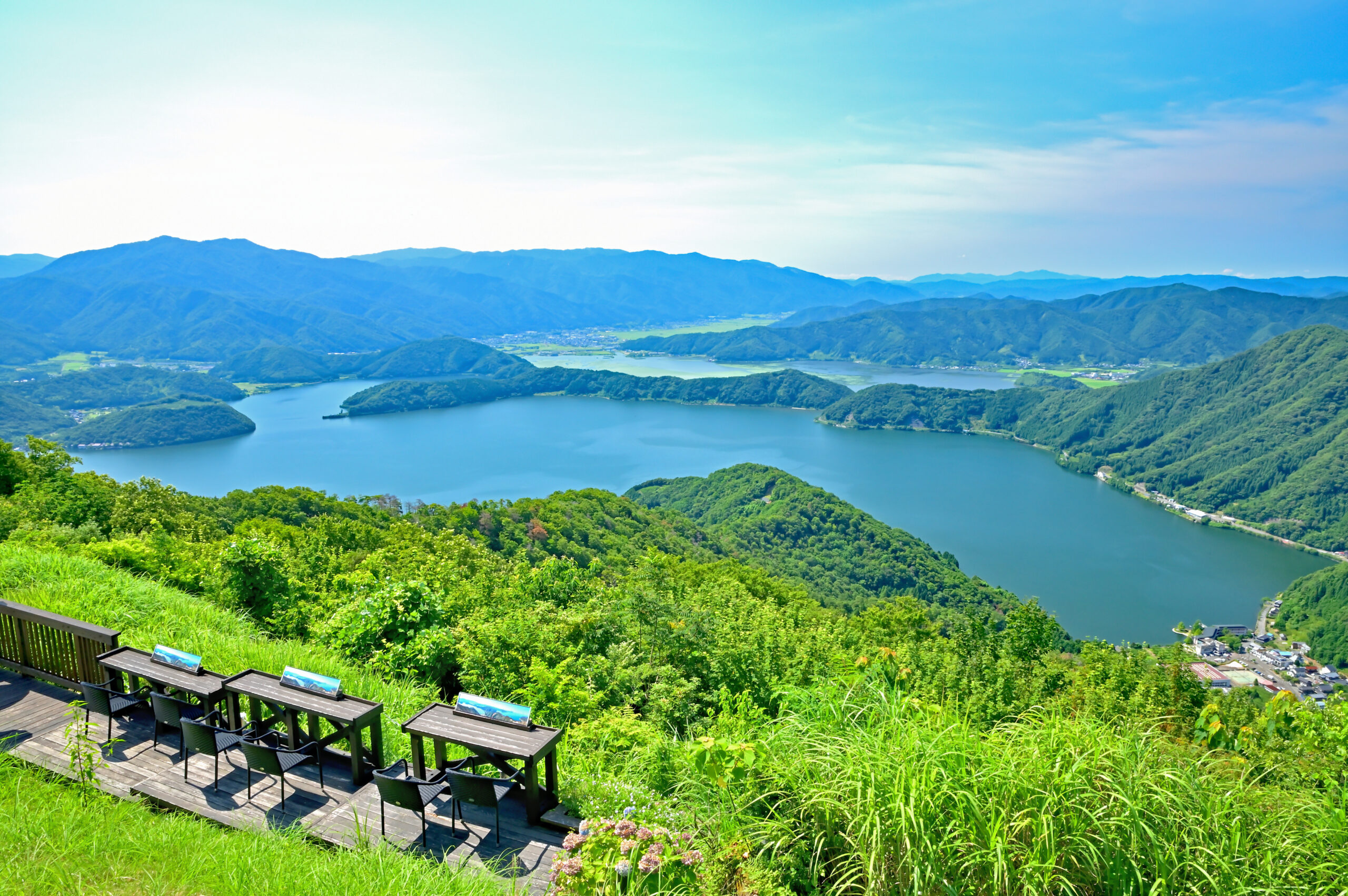 Terraced viewpoint overlooking a lake surrounded by green mountains, with a wooden bench railing in the foreground.