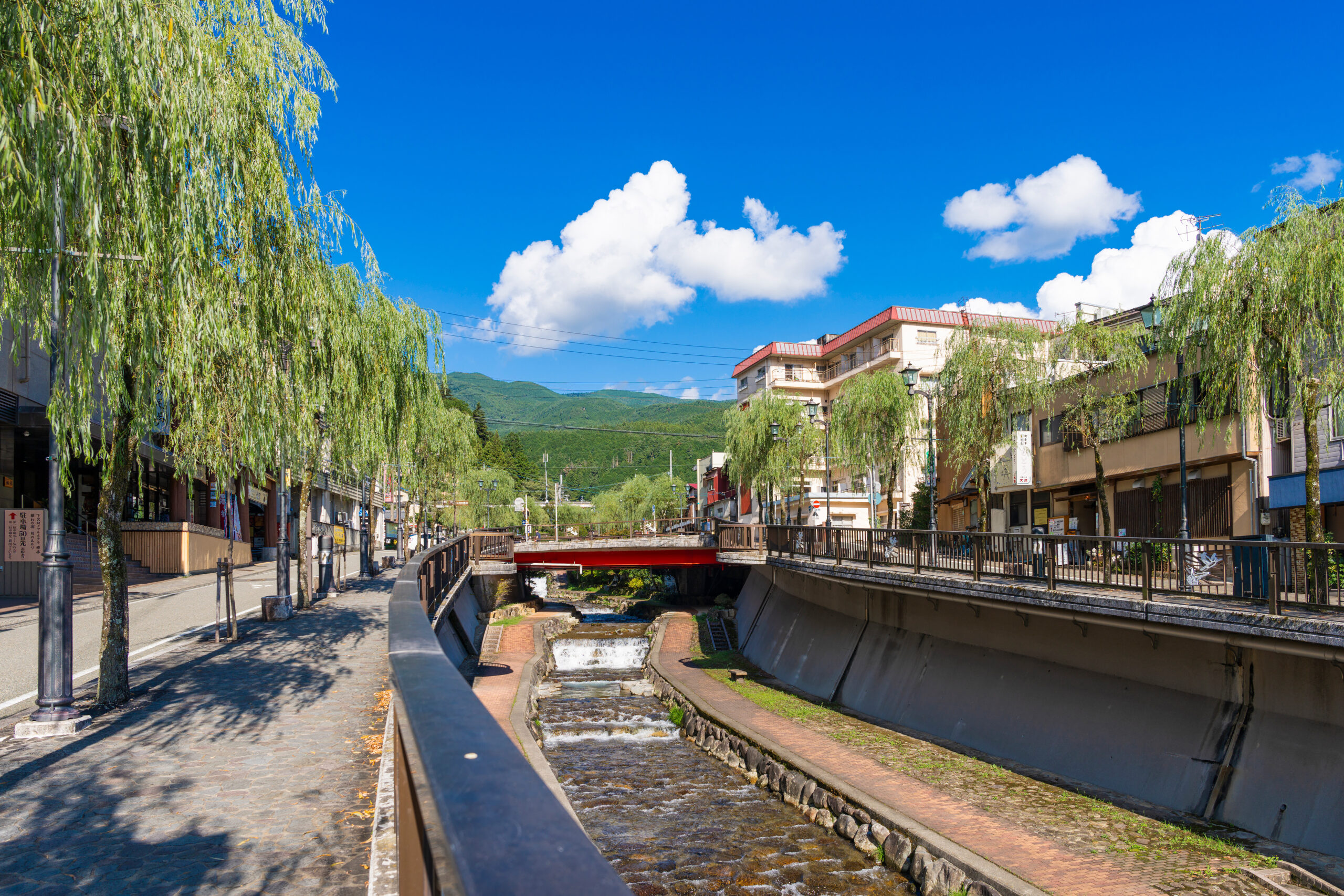 Sunny urban canal street lined with weeping willow trees, boats, and buildings against a bright blue sky with white clouds and distant mountains.
