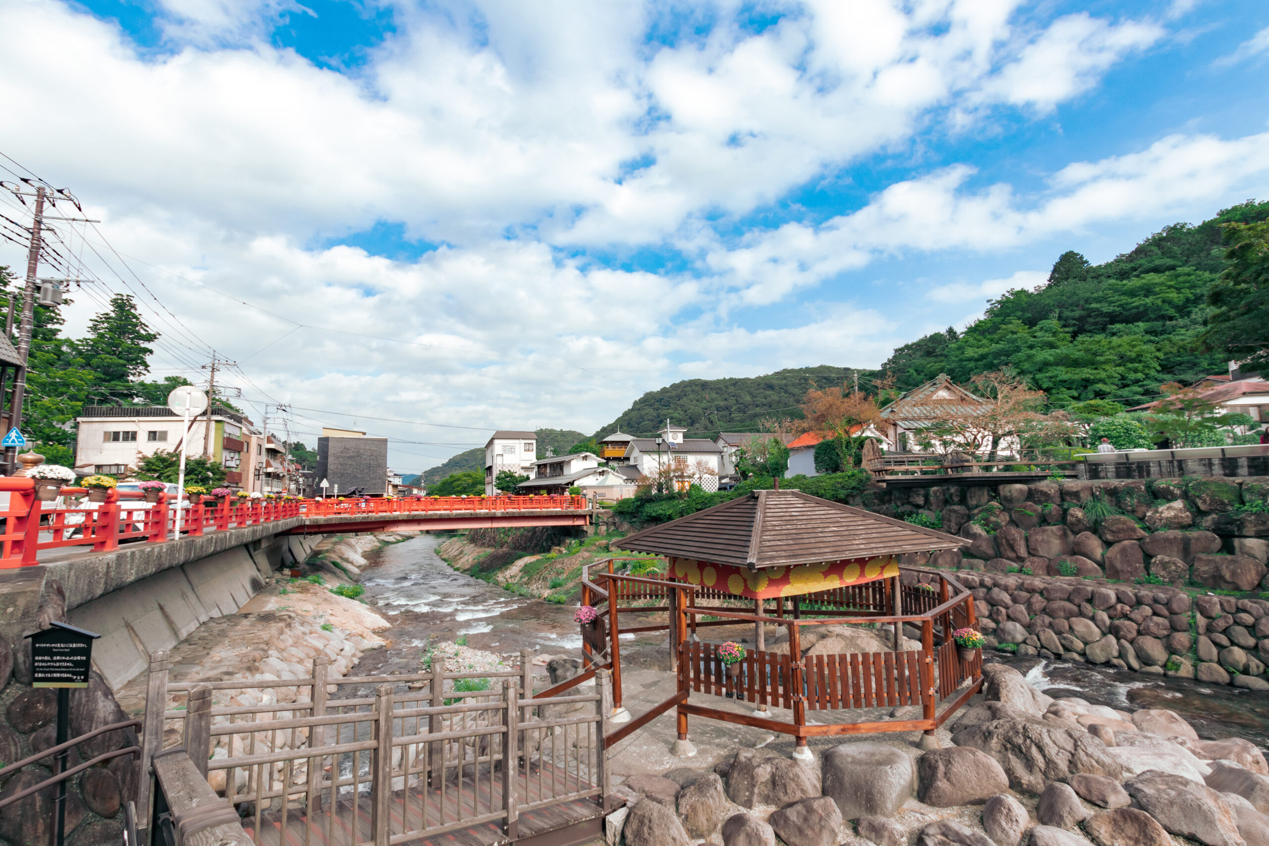 Riverside town with a red railing bridge, wooden walkways, and rocky riverbanks under a bright, partly cloudy sky.